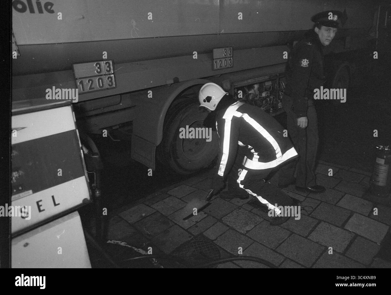 Perdita in petroliera, Julianalaan, incidenti, collisioni, incidenti, Haarlem, Julianalaan, Paesi Bassi, 27-11-1989 Whizgle News, Dutch Desk, i Paesi Bassi, 1950-2000 Un agente di polizia sta di guardia mentre un altro individuo, indossando un abito riflettente, ispeziona attentamente l'area vicino alle gomme di un camion. È visibile un estintore esterno, che suggerisce di prestare attenzione alla sicurezza durante l'ispezione. Foto Stock