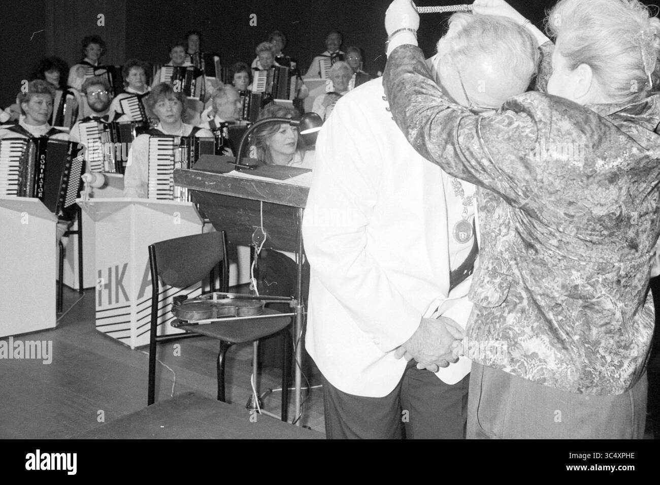 Joseph Stanek [Sindaco di SV@@tlá nad Sázavou, Repubblica Ceca] + Sindaco Schmitz, 24-11-1990 Whizgle News, Dutch Desk, Paesi Bassi, 1950-2000 Una donna posa cerimonialmente una medaglia intorno al collo di un uomo sul palco, mentre un gruppo di musicisti che suonano fisarmoniche osserva il momento con sorrisi e applausi. L'ambientazione suggerisce un evento celebrativo o di riconoscimento, mettendo in risalto l'amore condiviso per la musica e la comunità. Foto Stock