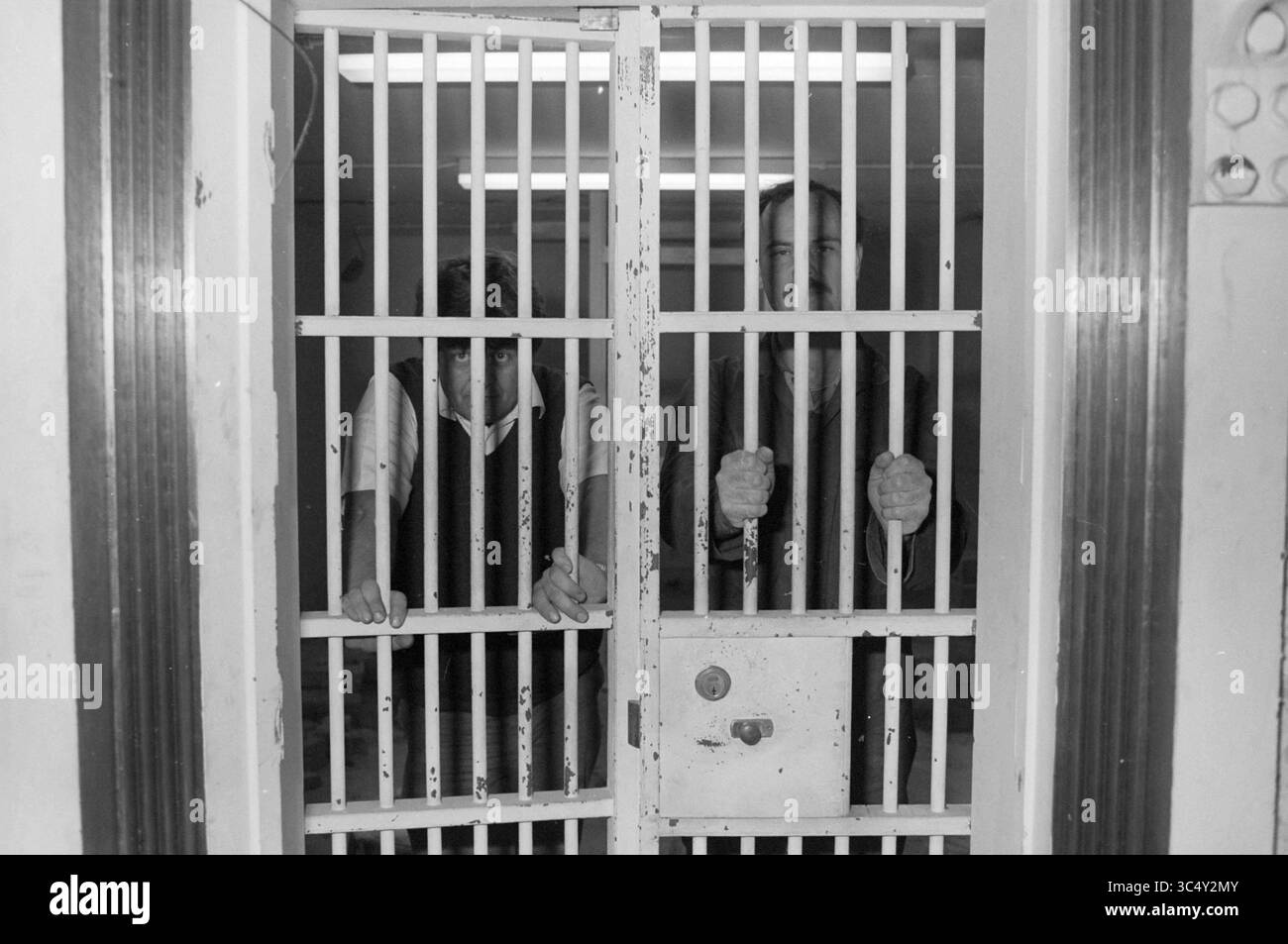 Due uomini dietro le sbarre, 00-00-1985 Whizgle News, Dutch Desk, Paesi Bassi, 1950-2000 due persone stanno dietro le sbarre della prigione, con uno che appare più animato e l'altro che sembra pensivo. L'ambiente duro enfatizza il loro confinamento, mentre il contrasto nelle espressioni suggerisce emozioni diverse sulla loro situazione. Foto Stock