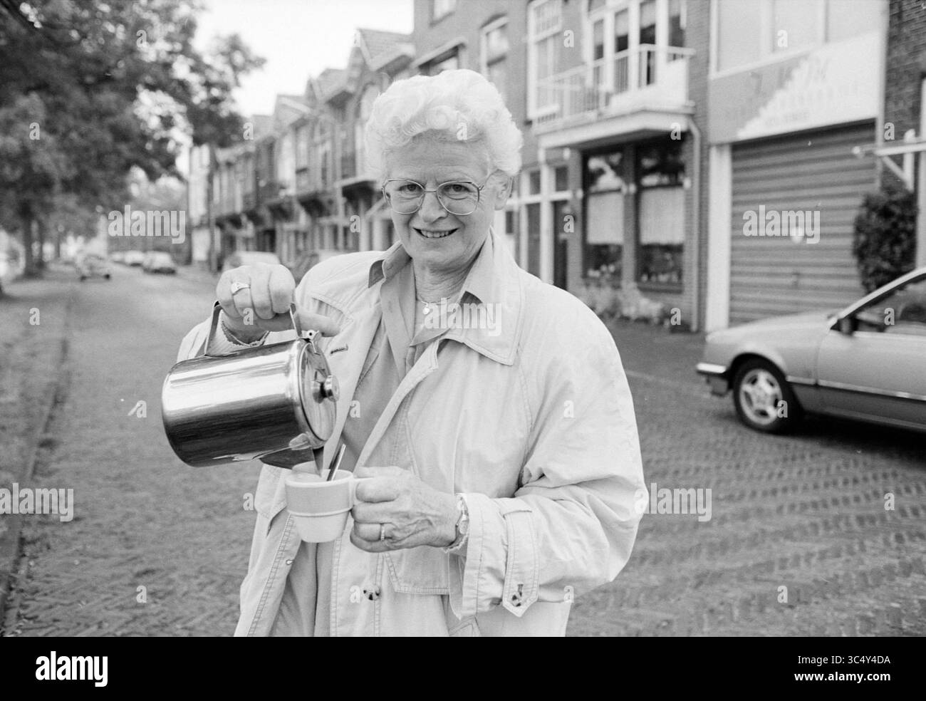Mrs. Van Sommeren, caffetteria, 15-06-1993 Whizgle News, Dutch Desk, Paesi Bassi, 1950-2000 una donna anziana con capelli bianchi ricci sorridente mentre versa il tè da un bollitore tradizionale in una tazza, in piedi su una strada affascinante fiancheggiata da case pittoresche. Foto Stock