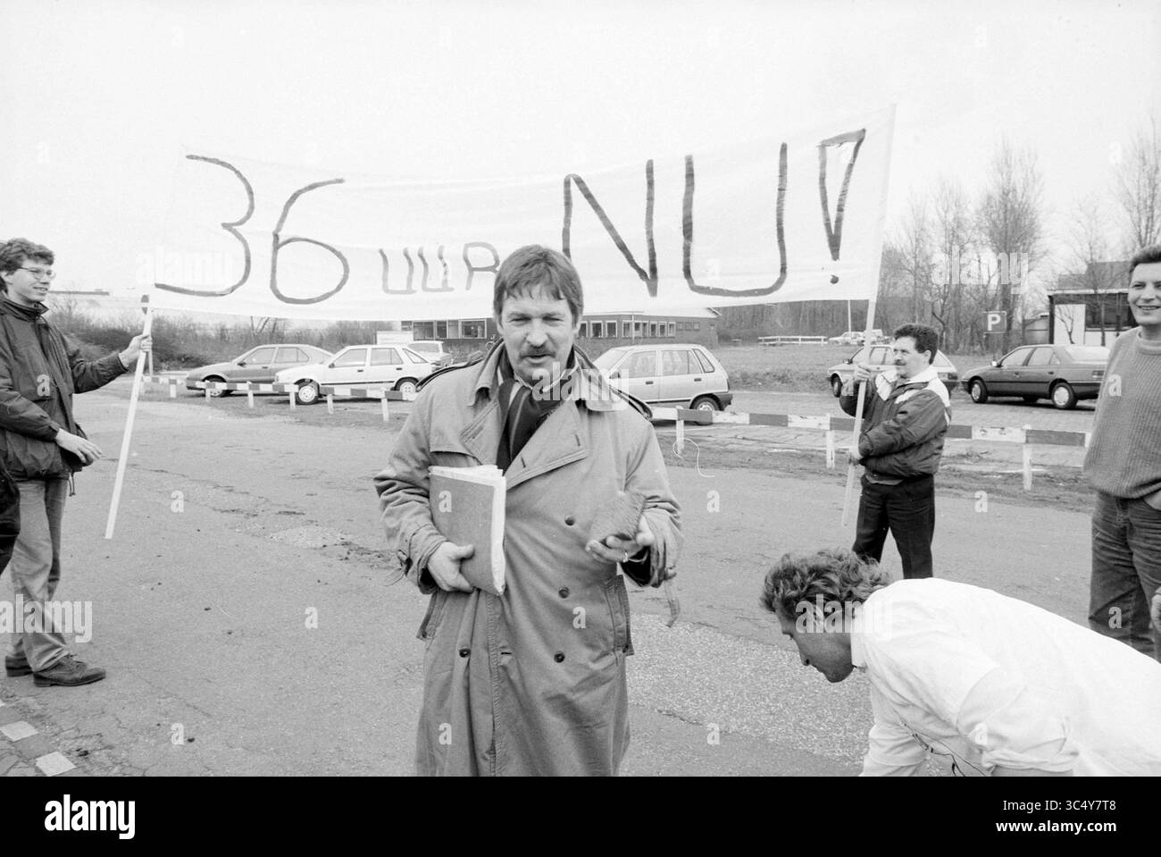 Azione "36 ore ora" dell'FNV presso gli Hoogovens, 20-03-1991 Whizgle News, Dutch Desk, Paesi Bassi, 1950-2000 Un gruppo di manifestanti è riunito lungo una strada, tenendo un grande striscione con testo audace che chiede azione. Un uomo sta in prima linea, indossando un trench e tenendo un libro, mentre sembra rivolgersi alla folla. Sullo sfondo, diverse auto sono parcheggiate, e alcuni individui sono impegnati in conversazione, evidenziando il loro sforzo collettivo e la loro determinazione. Foto Stock
