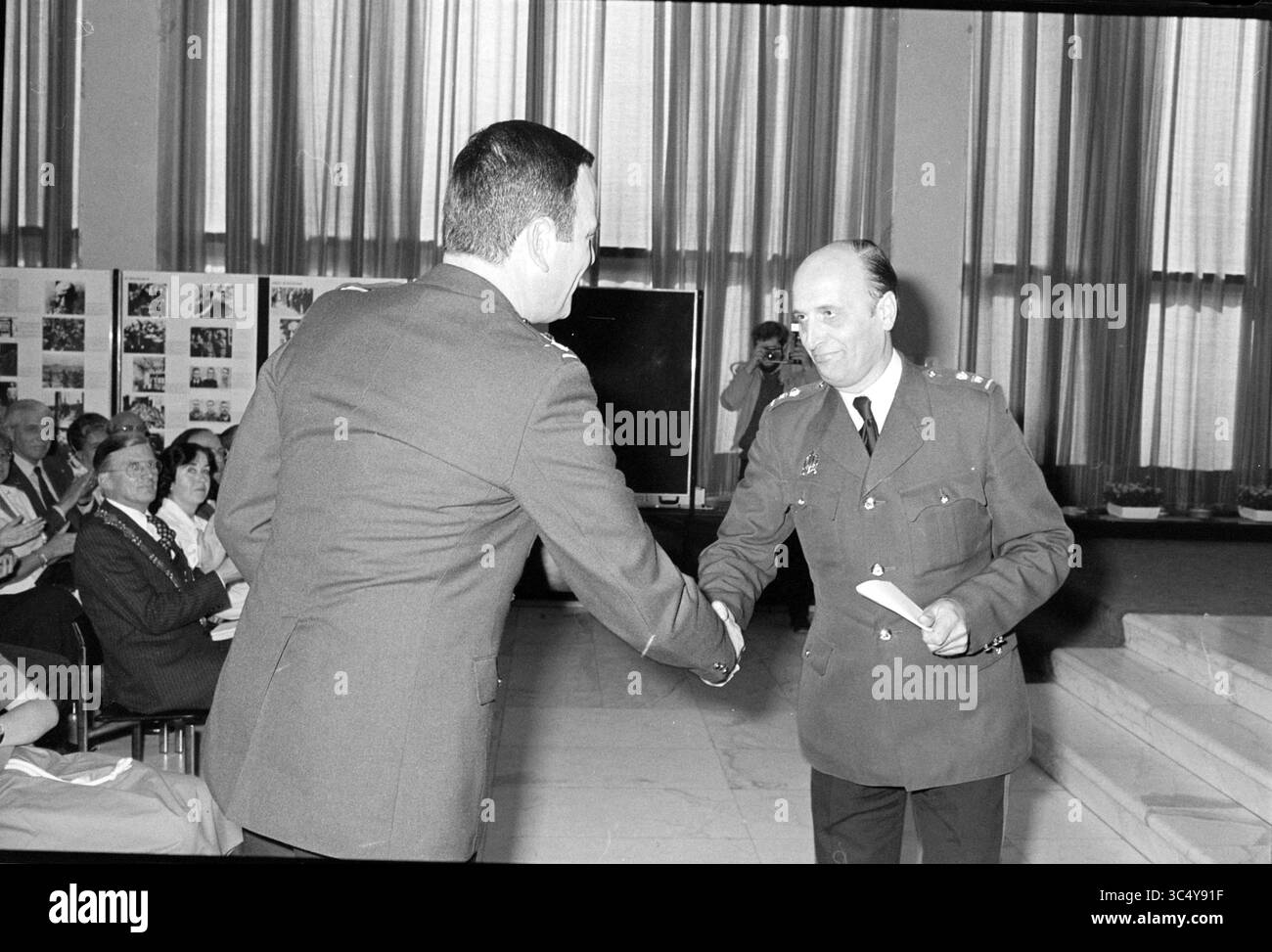 COM. Installazione Miller, polizia, 02-05-1980 Whizgle News, Dutch Desk, Paesi Bassi, 1950-2000 due individui sono impegnati in una stretta di mano durante un evento formale. Una persona, vestita in abbigliamento militare, sembra presentare qualcosa, possibilmente un certificato o un premio, all'altra, che è anche in abbigliamento formale. Sullo sfondo, un pubblico è seduto, osservando l'interazione, con esposizioni visive o fotografie disposte sulle pareti. L'ambientazione suggerisce una cerimonia o un evento di riconoscimento. Foto Stock