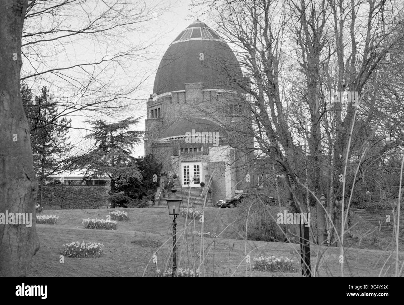 Exterior Crematorium Westerveld, Driehuis, 12-04-1995 Whizgle News, Dutch Desk, Paesi Bassi, 1950-2000 Un suggestivo edificio osservatorio con una caratteristica cupola si erge in modo prominente tra alberi senza foglie, circondato da fiori fioriti e un lampione d'epoca. Il paesaggio sereno crea uno stato d'animo contemplativo, evocando un senso di curiosità sull'universo. Foto Stock