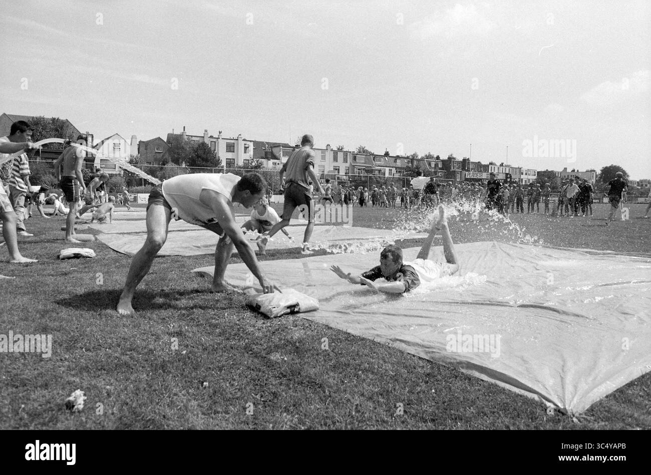 Children Practice Sliding, Kinheim, 06-08-1991 Whizgle News, Dutch Desk, Paesi Bassi, 1950-2000 Una vivace scena all'aperto cattura le persone che si godono una giornata estiva, con individui che scivolano su una superficie coperta d'acqua mentre altri tifanno da contorno. Risate e spruzzi riempiono l'aria mentre i partecipanti si impegnano in una competizione giocosa, creando un'atmosfera divertente ed energica. Foto Stock
