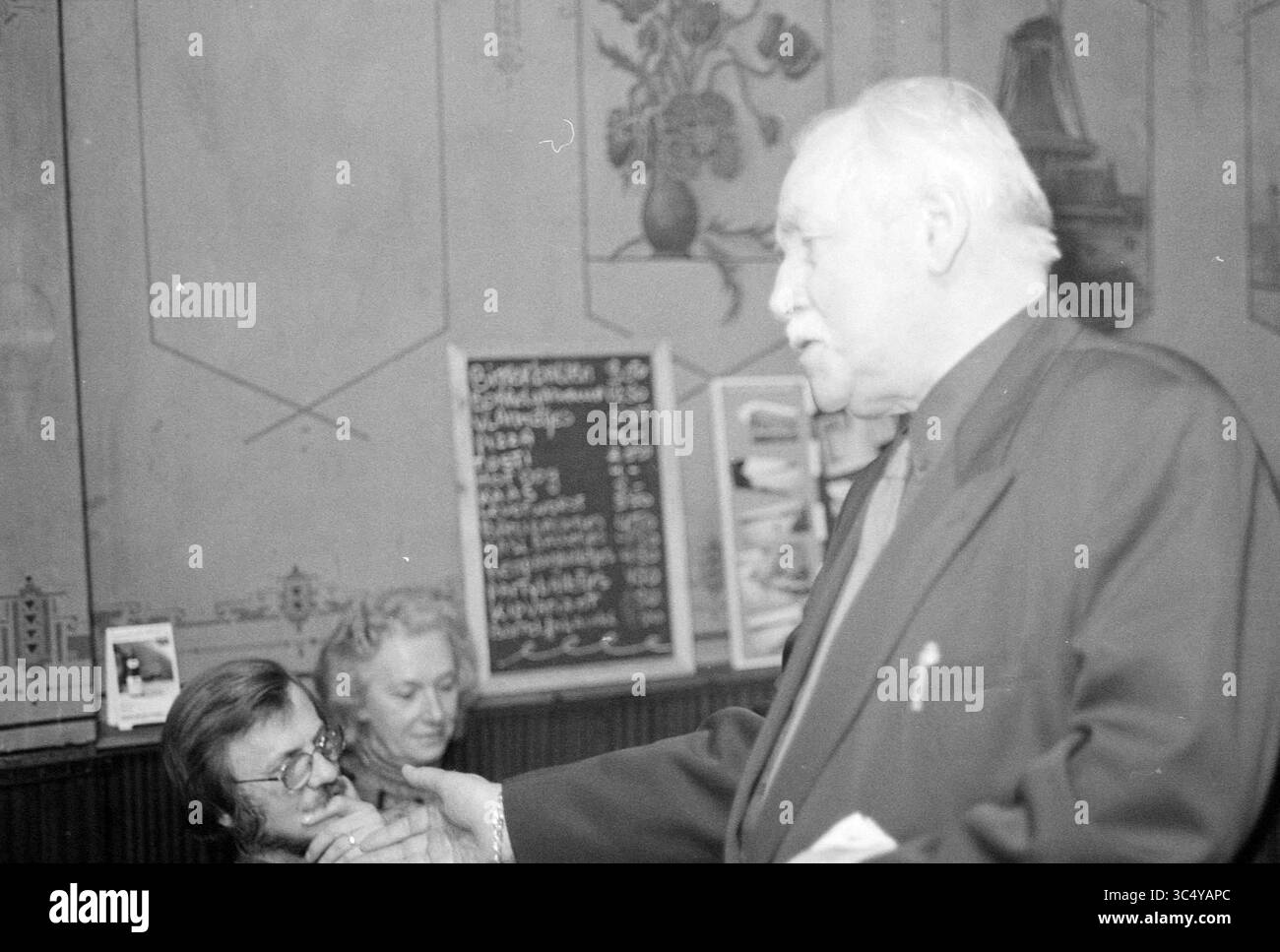 Jaap v.d. Tear, 15-02-1994 Whizgle News, Dutch Desk, Paesi Bassi, 1950-2000 Un uomo in tuta gesti mentre parla, con una coppia seduta vicino che appare impegnata in conversazione. Lo sfondo presenta pareti decorative e un menu che contribuisce a creare un'atmosfera calda e sociale. Foto Stock