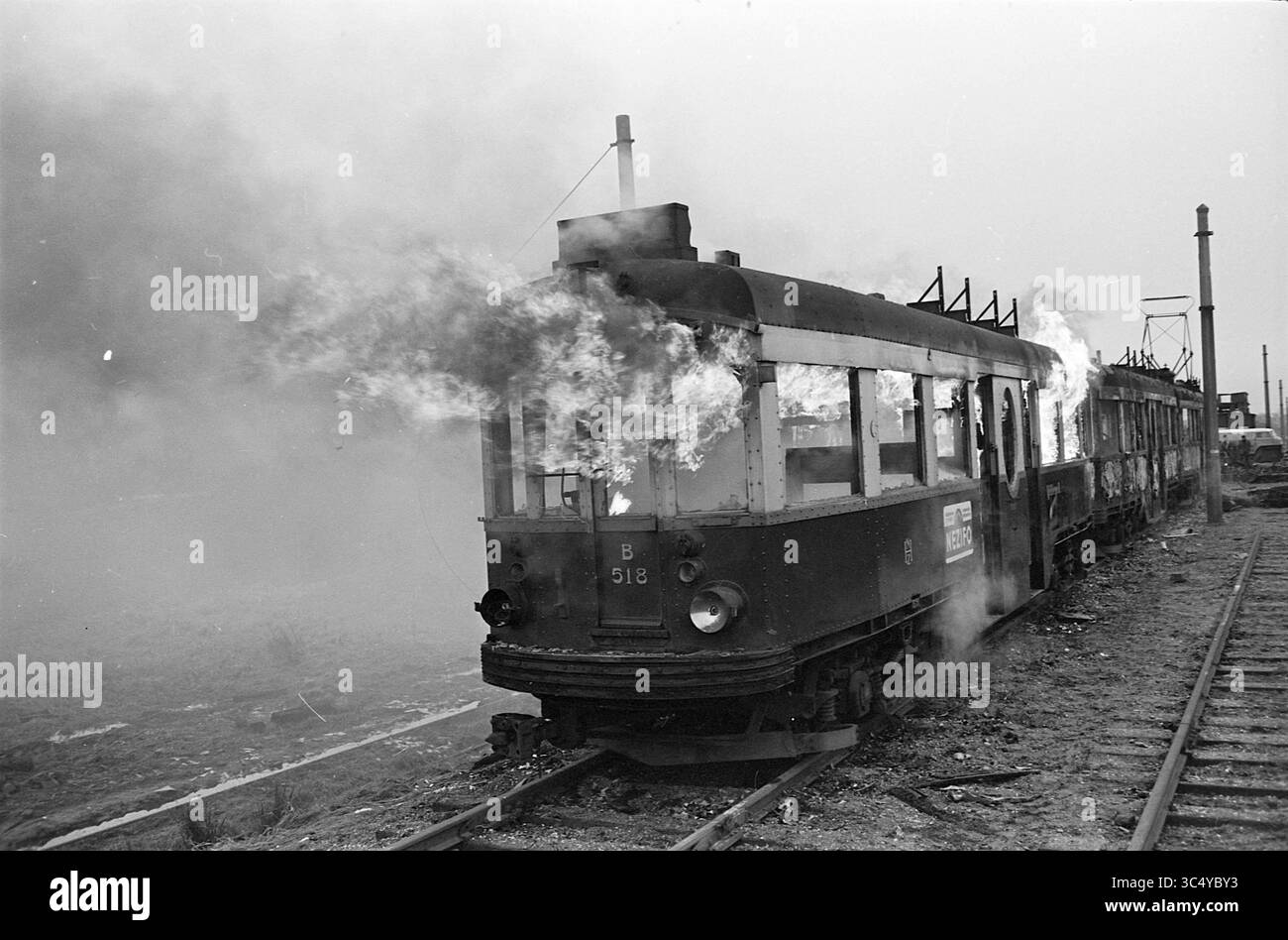 Demolizione di attrezzature NZH. I tram sono stati incendiati, lasciando solo il metallo a cui gli operai della demolizione erano interessati. Ecco come i tram blu finirono ad Aldegeest, un sito tra Voorschoten e Leiden, vicino al Korte Vliet, incendi, vigili del fuoco, Voorschoten, 14-12-1961 Whizgle News, Dutch Desk, Paesi Bassi, 1950-2000 Un vagone ferroviario inondato di fiamme, fumo che si diffonde nell'aria mentre si siede sui binari, con resti della sua struttura visibili in mezzo alla distruzione dell'incendio. Foto Stock