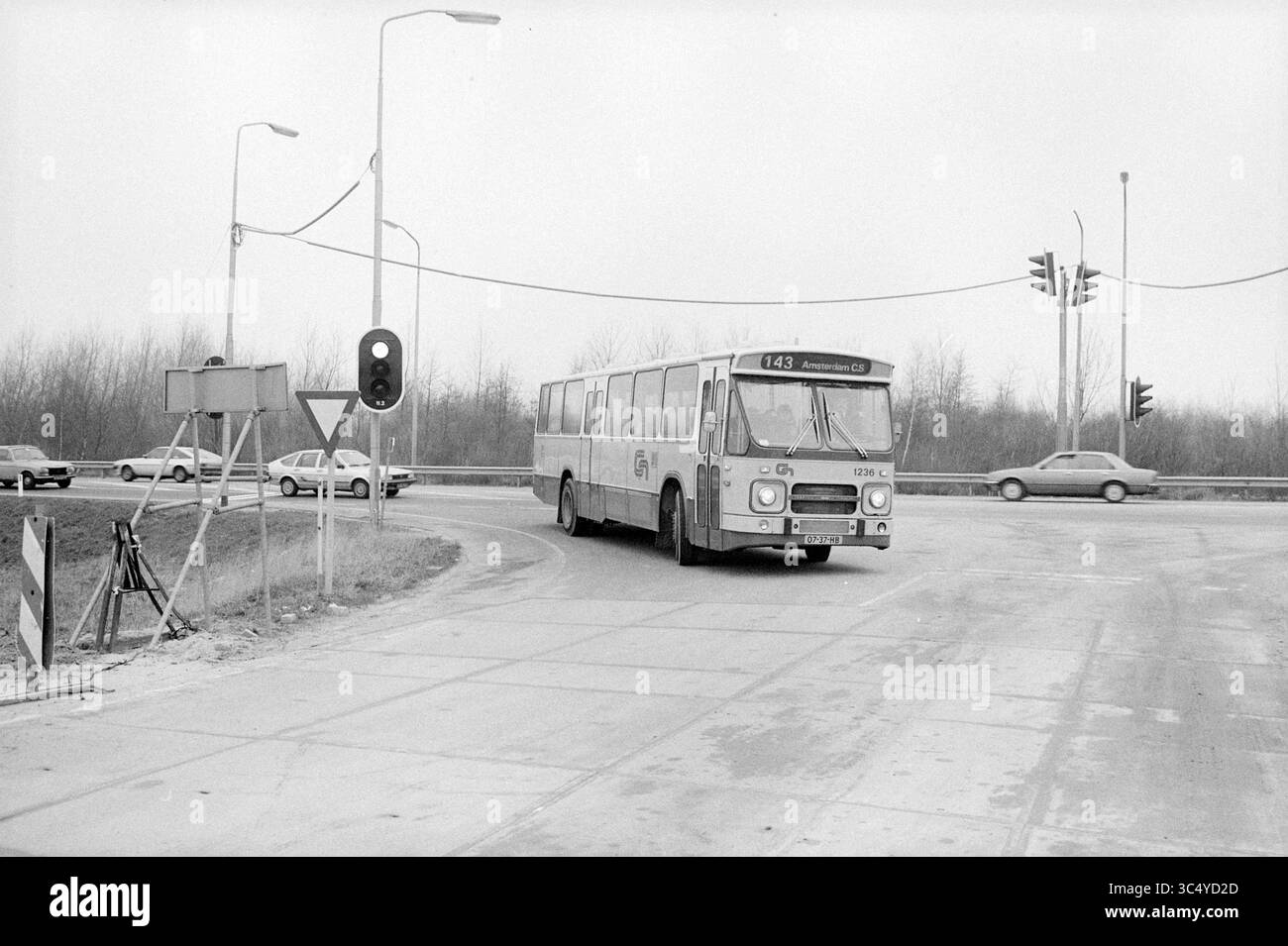 Nuova linea di autobus, Overbos, Hoofddorp, autobus, tour in autobus, Noord Zuid Hollandse Vervoersmaatschappij N.V., nuova Zelanda, Hoofddorp, Paesi Bassi, 06-03-1984 Whizgle News, Dutch Desk, Paesi Bassi, 1950-2000 Un autobus cittadino fa una svolta a una rotatoria, avvicinandosi a un segnale stradale, mentre alcune auto vi attendono dietro. Il paesaggio circostante è sparso, con alberi sullo sfondo che lasciano intendere un ambiente tranquillo. Foto Stock