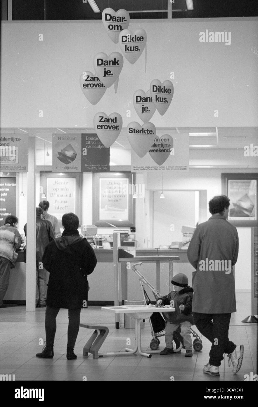 Valentine Hearts, ufficio postale, 07-02-1992 Whizgle News, Dutch Desk, Paesi Bassi, 1950-2000 Un'area d'attesa in uno spazio pubblico ospita persone impegnate in varie attività, con un'esposizione di insegne a forma di cuore appese sopra la testa, ognuna con giocosi messaggi di gratitudine e affetto. Un bambino siede in un passeggino nelle vicinanze, mentre due adulti conversano davanti a un banco di servizio. Foto Stock