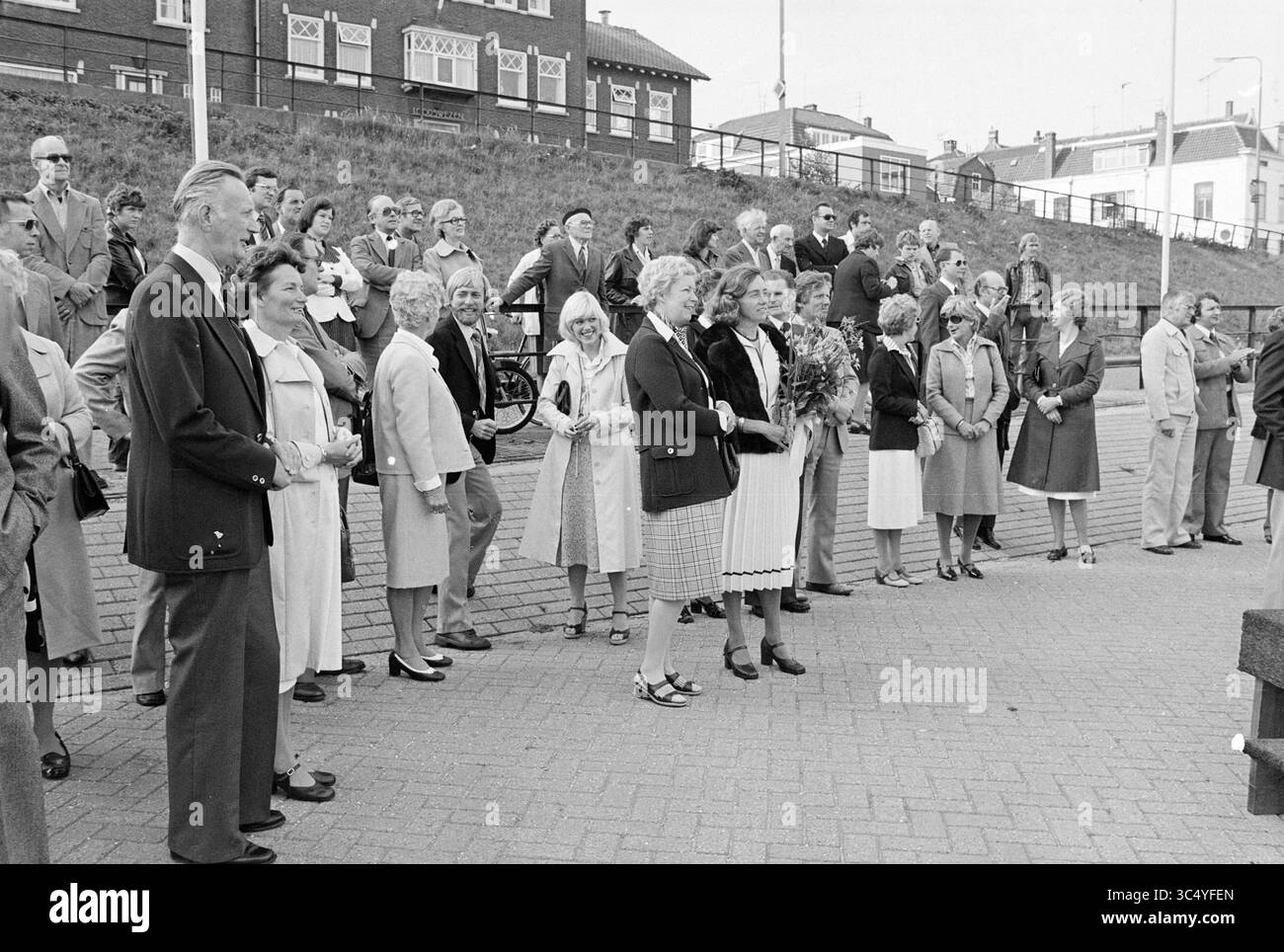 Trasferimento della gara d'appalto pilota The Pluvier, consegna e trasferimenti, 26-08-1977 Whizgle News, Dutch Desk, Paesi Bassi, 1950-2000 Un grande gruppo di persone si radunò all'aperto, indossando una varietà di abbigliamento alla moda, sorridendo e interagendo. Alcuni tengono bouquet, suggerendo un'occasione celebrativa, mentre altri si trovano casualmente in una formazione rilassata, creando un senso di comunità e coinvolgimento. Edifici e un'area erbosa fanno da sfondo. Foto Stock