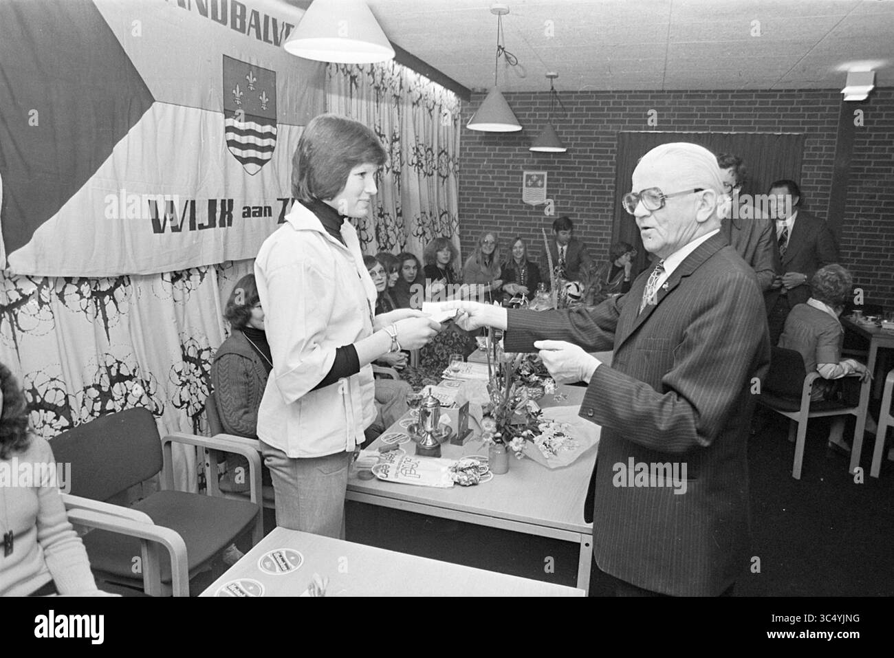 Ricevimento campione di pallamano squadra Wijk aan Zee, pallamano, ricevimenti, 14-03-1975 Whizgle News, Dutch Desk, i Paesi Bassi, 1950-2000 Un evento formale si svolge in una sala comune, dove un uomo anziano con una tuta presenta un premio o un certificato a una giovane donna con una giacca bianca. Sullo sfondo, diverse persone sono sedute a tavoli adornati da decorazioni, con striscioni che espongono insegne locali appese alle pareti. L'atmosfera suggerisce una celebrazione o un riconoscimento del successo all'interno della comunità. Foto Stock