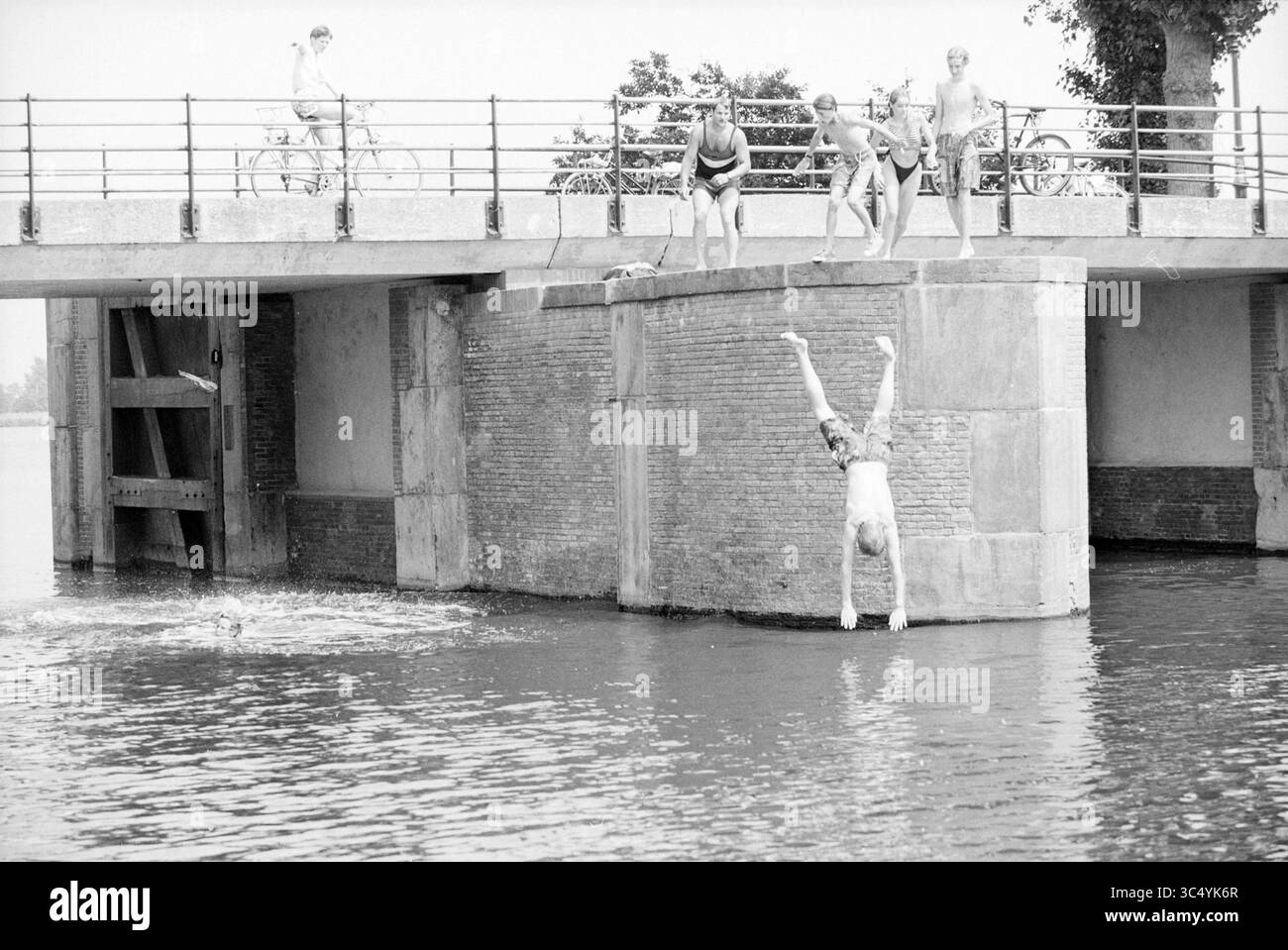 Nuoto divertente da un ponte vicino all'acqua, 00-06-1992 Whizgle News, Dutch Desk, Paesi Bassi, 1950-2000 Un gruppo di giovani nuotatori gode di una giornata di sole, con una persona che si tuffa in acqua mentre altri tifo dal ponte sopra. Nelle vicinanze, un ciclista si ferma per guardare, creando una vivace scena di divertimento estivo e cameratismo. Foto Stock