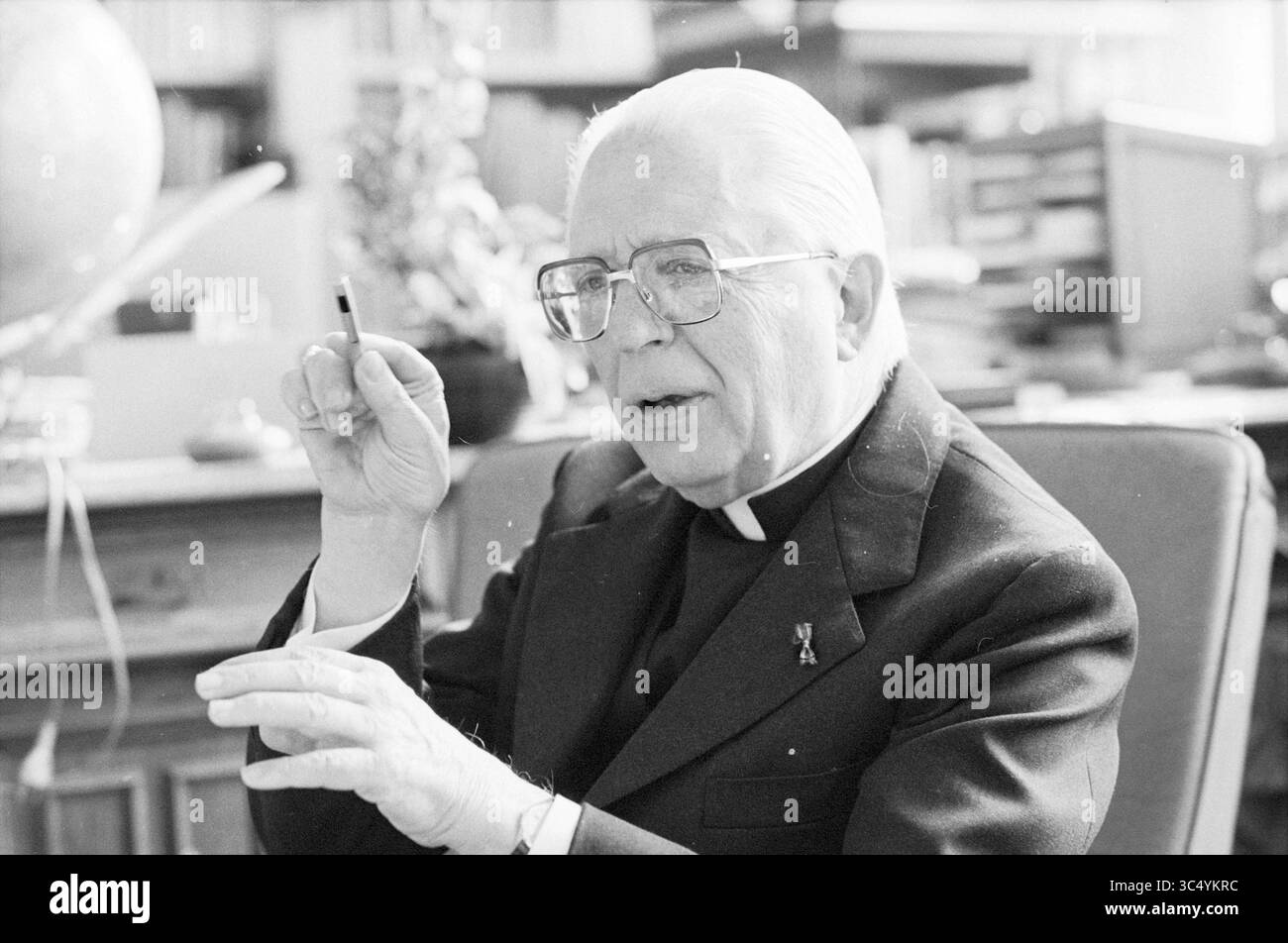Ritratto di Mgr. Zwartkruis, Persons R.K. and Protestant, 24-03-1982 Whizgle News, Dutch Desk, Paesi Bassi, 1950-2000 gesti Di Una figura premurosa con una penna mentre si siede in un ambiente accademico, circondato da scaffali di libri e materiali accademici. Sembra essere impegnato in una discussione o riflessione, mostrando una miscela di conoscenza e autorità. Foto Stock