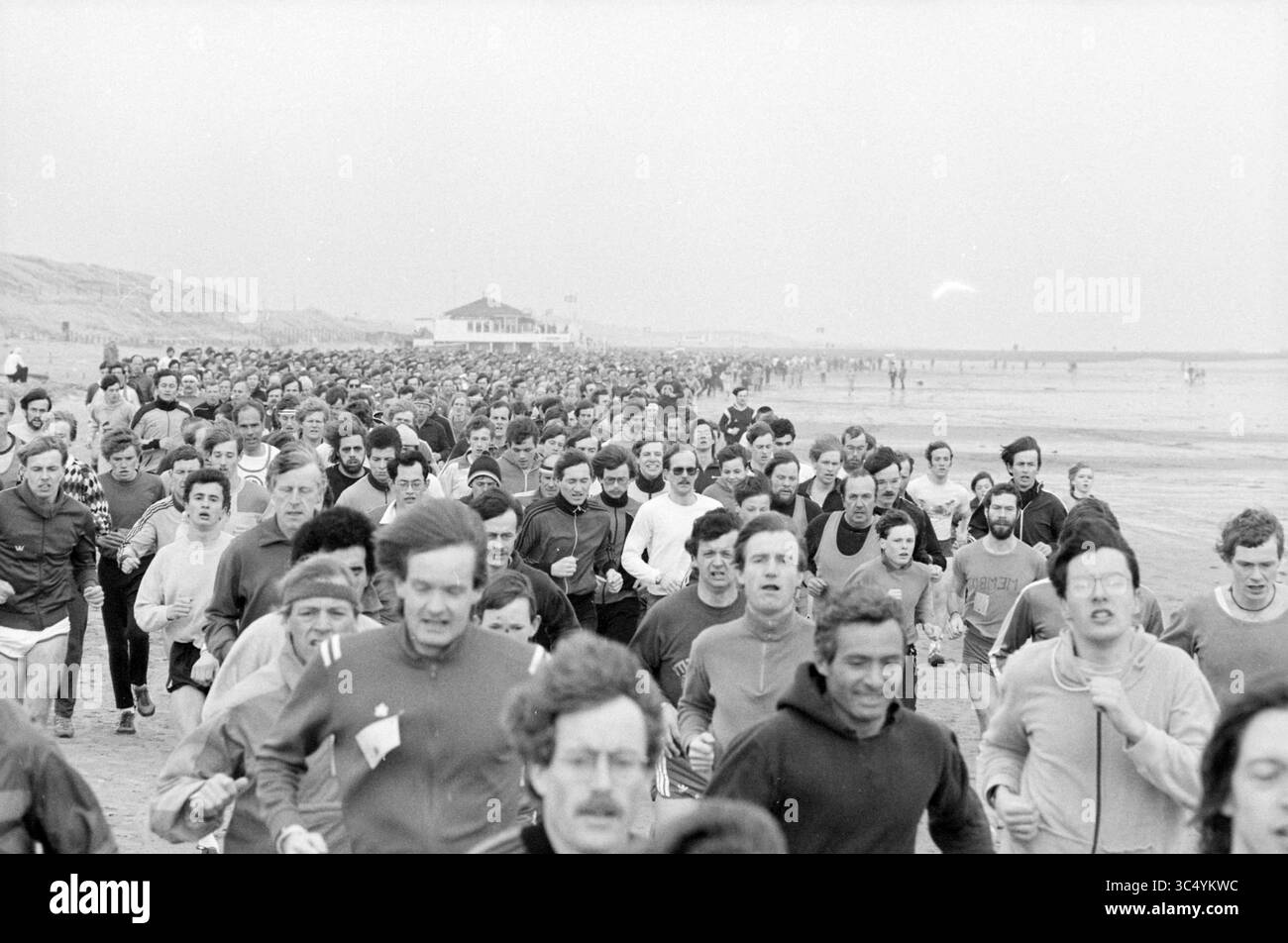 AAC Beach run, IJmuiden, running, IJmuiden, Paesi Bassi, 06-03-1983 Whizgle News, Dutch Desk, Paesi Bassi, 1950-2000 Una folla affollata di corridori carica lungo la costa, incarnando l'energia di un evento di massa. I volti spaziano dalla determinazione all'eccitazione, mentre i partecipanti, vestiti con vari outfit sportivi, si spingono in avanti insieme sullo sfondo di un cielo nuvoloso e di un molo lontano. Foto Stock