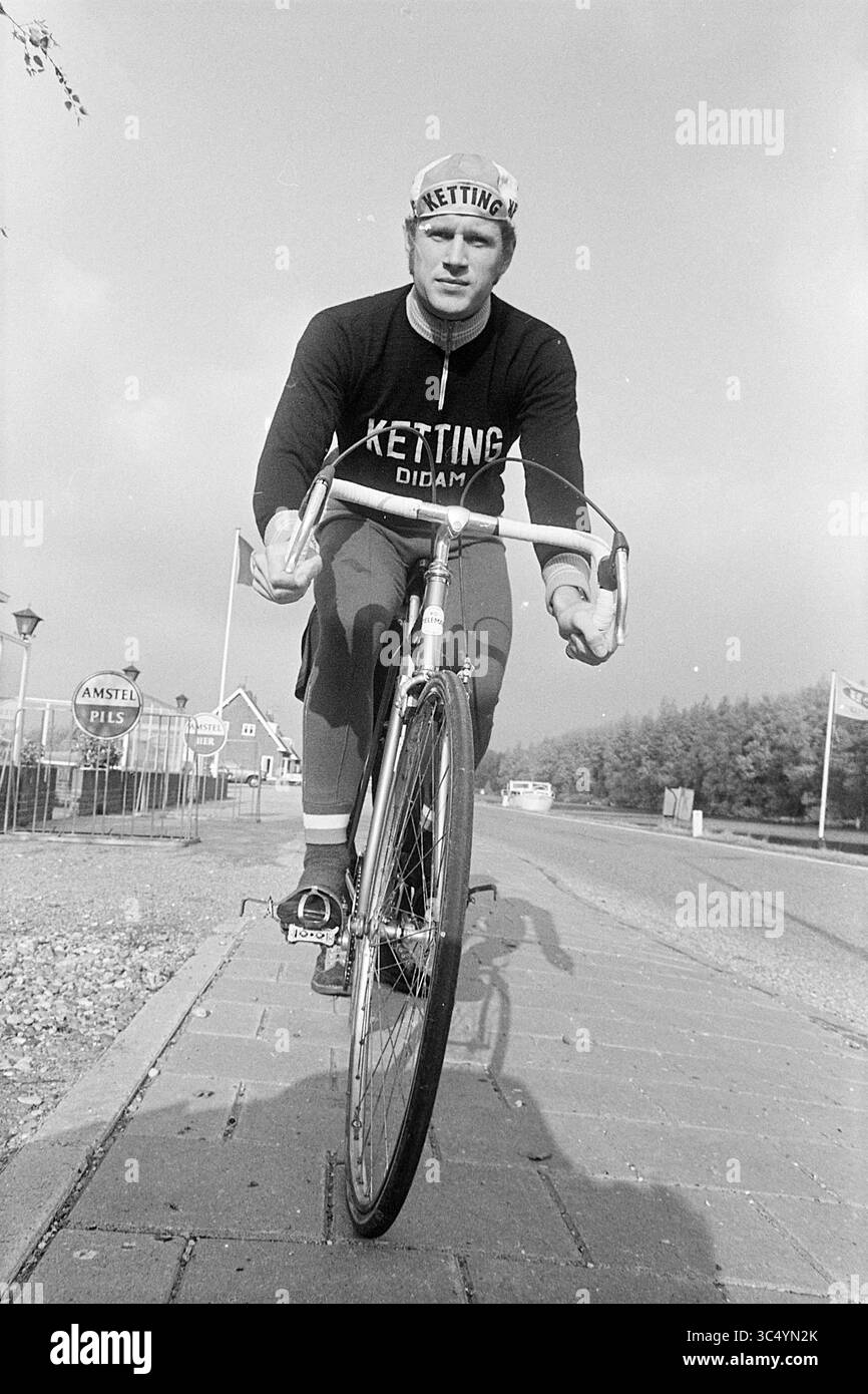 Cyclist Whizgle News, Dutch Desk, Paesi Bassi, 1950-2000 Un ciclista indossa una maglia e un berretto della squadra, appoggiandosi in avanti sulla propria bicicletta mentre percorre un percorso asfaltato. Le bandiere sono disposte sullo sfondo lungo la strada, a indicare un'atmosfera competitiva. Foto Stock