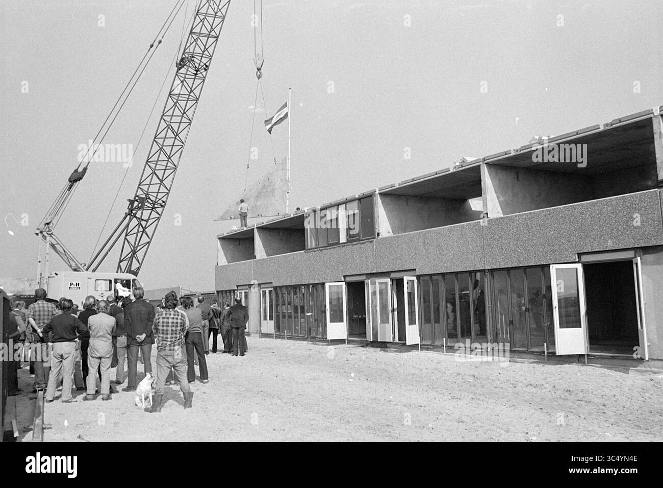 Le case di nuova costruzione raggiungeranno il punto più alto?, 21-12-1973 Whizgle News, Dutch Desk, Paesi Bassi, 1950-2000 Un gruppo di persone si riunisce di fronte a un edificio parzialmente costruito, mentre una gru solleva una bandiera. L'ambiente sembra essere un cantiere, con l'attenzione sul design architettonico unico dell'edificio e la bandiera che viene issata. Foto Stock