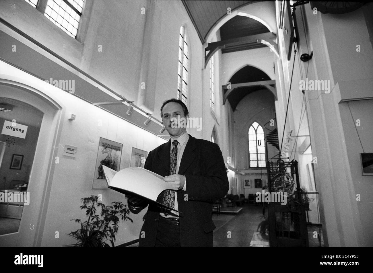 Interno degli archivi comunali, Jansstraat 40, Haarlem, Jansstraat, Nederland, 29-01-1996 Whizgle News, Dutch Desk, Paesi Bassi, 1950-2000 Un uomo in abbigliamento formale si erge in un ampio interno a volta, tenendo in mano un grande libro mentre guarda con attenzione lo spettatore. Lo spazio presenta alte finestre e una serie di sedie sullo sfondo, suggerendo un ambiente incentrato sull'arte e l'impegno della comunità. Foto Stock