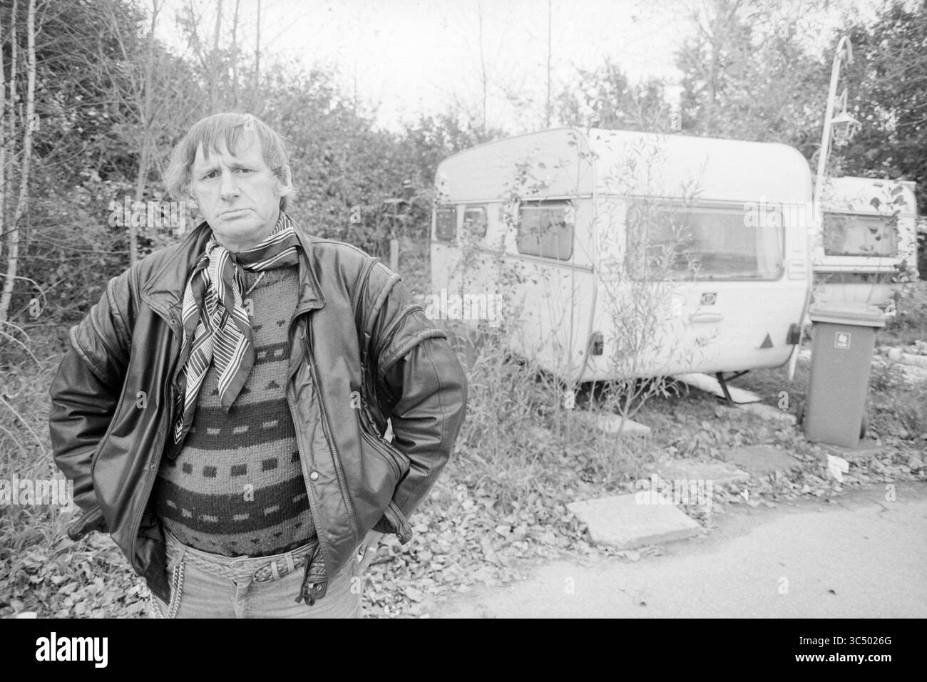 Sjors la Moine, campo carovaniere, IJmuiden, IJmuiden, Paesi Bassi, 06-10-1993 Whizgle News, Dutch Desk, Paesi Bassi, 1950-2000 Un uomo si trova con fiducia di fronte a una carovana d'epoca in mezzo alla vegetazione ricoperta, indossando una giacca di pelle e un maglione a fantasia, con un'espressione seria che suggerisce introspezione o contemplazione. Foto Stock