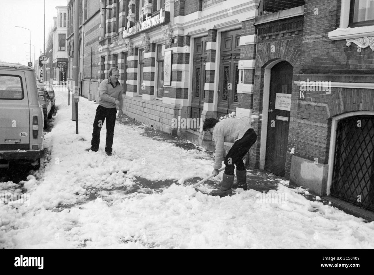 Clearing Snow for business, Snow, 24-01-1984 Whizgle News, Dutch Desk, Paesi Bassi, 1950-2000 due uomini lavorano insieme per scacciare la neve da un marciapiede, con uno in piedi e supervisionando mentre l'altro si piega verso il basso per liberare la neve. Sullo sfondo, un edificio caratterizzato da mattoni ornati e grandi finestre aggiunge carattere alla scena, mentre un veicolo parcheggiato è visibile nelle vicinanze. Foto Stock