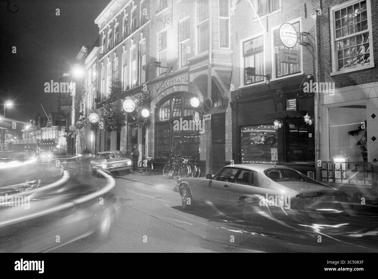 Evening Atmosphere River Fish Market and pubs, Haarlem, Riviervismarkt, Paesi Bassi, 21-12-1992 Whizgle News, Dutch Desk, Paesi Bassi, 1950-2000 Una vivace scena di strada di notte con vetrine illuminate, auto d'epoca parcheggiate lungo il marciapiede e movimento sfocato dei veicoli di passaggio che creano un senso di movimento e attività. Le lampade da strada emettono un caldo bagliore, evidenziando l'affascinante architettura e l'atmosfera invitante. Foto Stock