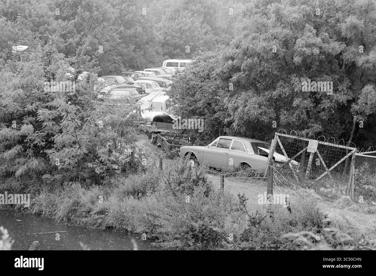 Cantiere di rottami Garenkokerskade, demolizione, demolizione, cantieri di rottami, Haarlem, Garenkokerskade, Paesi Bassi, 09-06-1977 Whizgle News, Dutch Desk, Paesi Bassi, 1950-2000 Una recinzione fatiscente confina con un corso d'acqua, con fogliame sovrastato e una collezione di auto abbandonate parzialmente nascoste sullo sfondo. Un veicolo d'epoca è parcheggiato vicino al bordo, suggerendo un'area un tempo trafficata ora lasciata all'invasione della natura. Foto Stock