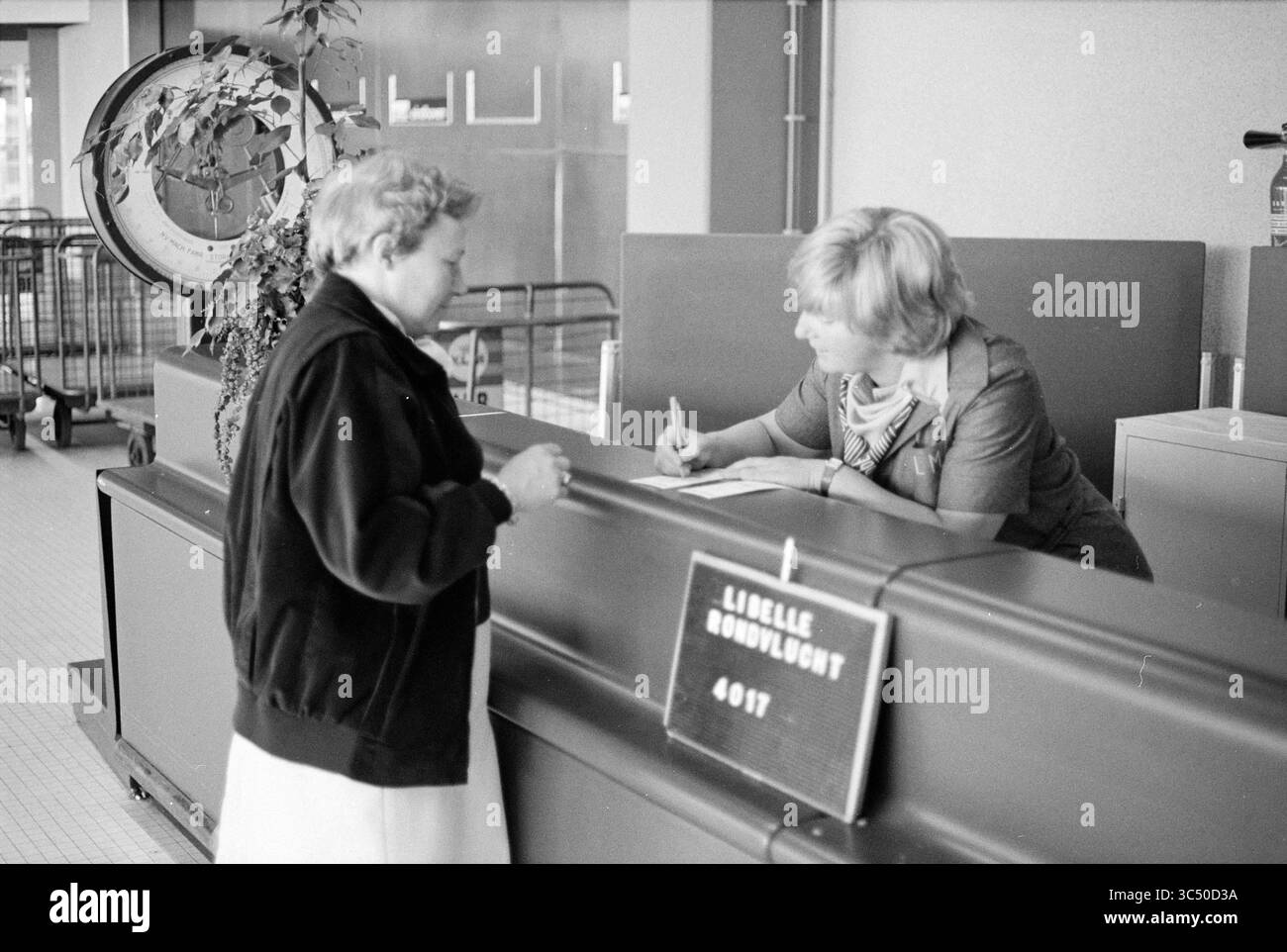 Volo Dragonfly, crociera, voli turistici, voli turistici, 01-06-1979 Whizgle News, Dutch Desk, Paesi Bassi, 1950-2000 Una donna si trova al banco della reception, impegnata in una conversazione con un membro del personale che prende appunti. Dietro di loro, un grande orologio e vari segni contribuiscono all'atmosfera affollata dello spazio. Foto Stock