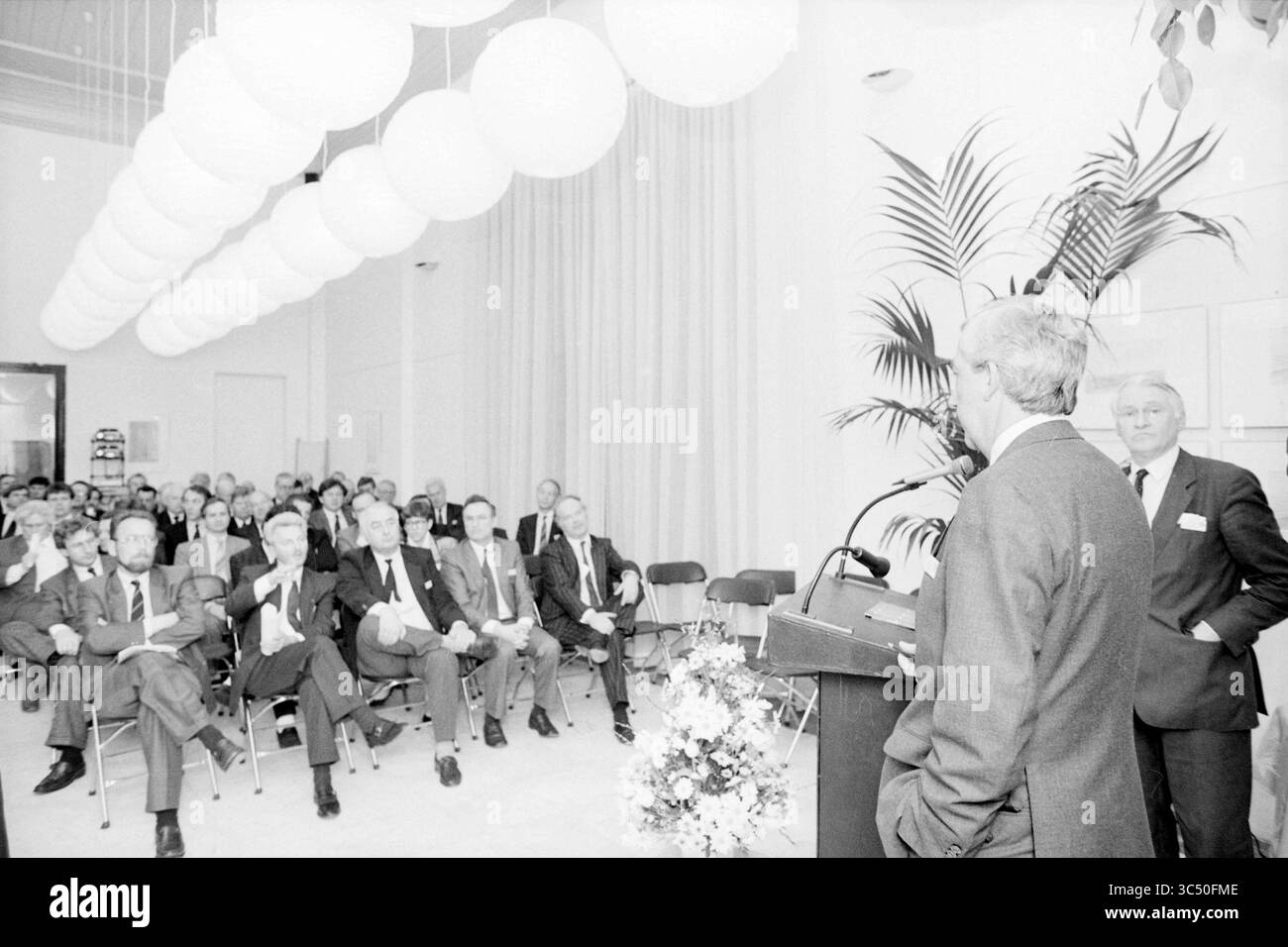 Incontro degli imprenditori della camera di commercio con gemme. E prov., riunioni, camera di commercio e fabbriche, 10-03-1988 Whizgle News, Dutch Desk, Paesi Bassi, 1950-2000 Un oratore si trova su un podio rivolgendosi a un pubblico impegnato seduto in file, con espressioni attente e note occasionali prese. La sala è dotata di eleganti lampade rotonde e piante decorative, che creano un'atmosfera professionale per l'evento. Foto Stock