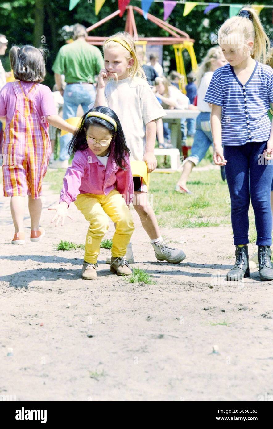 Marble Day Playground, Santpoort, Santpoort, 02-06-1998 Whizgle News, reception olandese i Paesi Bassi, 1950-2000 Un gruppo di bambini gioca all'aperto in un ambiente vivace, con una ragazza che si piega a terra, concentrandosi su qualcosa nello sporco, mentre altri si dedicano a varie attività nelle vicinanze. Decorazioni colorate suggeriscono un'atmosfera festosa. Foto Stock