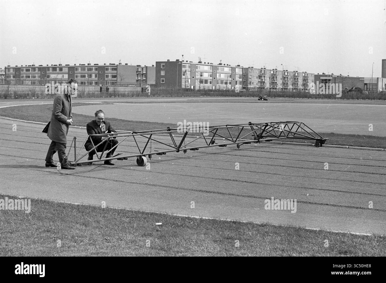 Nuova pista di cenere PIM Mulier Sports Park, settimana sportiva e sportiva, 16-04-1964 Whizgle News, Dutch Desk, Paesi Bassi, 1950-2000 due uomini sono impegnati in attività con una struttura lunga e leggera su una pista ampia e aperta. Sembra che stiano esaminando o regolando attentamente la struttura, che somiglia a un segmento di un meccanismo più grande. Sullo sfondo, ci sono diversi edifici, che suggeriscono un ambiente urbano. La scena trasmette un senso di sperimentazione o sforzo ingegneristico. Foto Stock
