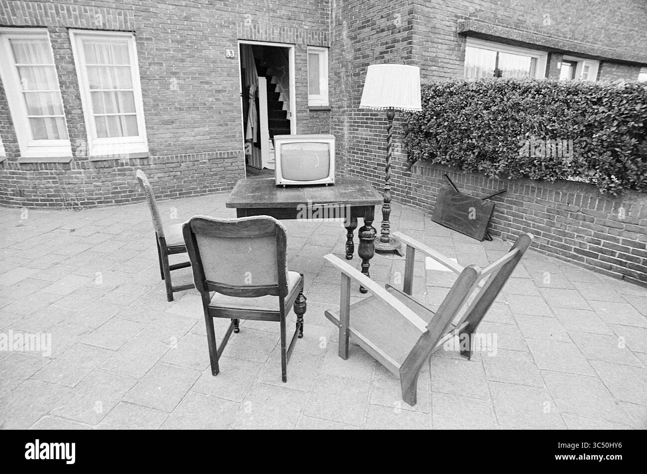 Contenuti esterni in Zandvoort, sfratto, 20-07-1973 Whizgle News, Dutch Desk, Paesi Bassi, 1950-2000 Un piccolo patio esterno presenta un tavolo di legno circondato da quattro sedie, con un vecchio televisore posizionato sul tavolo. Sullo sfondo, una porta porta conduce a un edificio e una lampada si trova nelle vicinanze, aggiungendo un tocco inaspettato alla scena. Una siepe delinea il bordo dell'area, contribuendo a creare un'atmosfera intima ma non convenzionale. Foto Stock