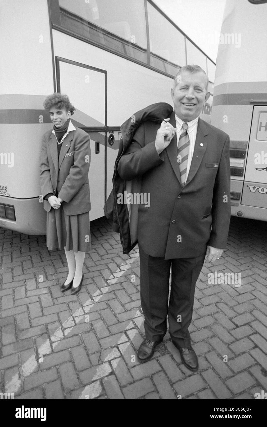 Nuovo staff NZH, 06-01-1992 Whizgle News, Dutch Desk, Paesi Bassi, 1950-2000 Un uomo con una tuta formale e cravatta si erge con sicurezza con un cappotto drappeggiato sulla spalla, sorridendo alla telecamera. Dietro di lui, una donna con un vestito coordinato appare leggermente riservata, accanto a un autobus in un parcheggio. Foto Stock
