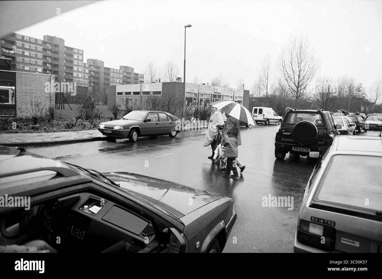 Padre del traffico, signora The Hope, 21-12-1995 Whizgle News, Dutch Desk, Paesi Bassi, 1950-2000 Una famiglia attraversa una strada bagnata, con una persona che tiene in mano un ombrello a righe. Nelle vicinanze, le auto parcheggiate fiancheggiano la strada, mentre sullo sfondo si possono vedere diversi edifici, leggermente oscurati da un'atmosfera nebbiosa. Foto Stock