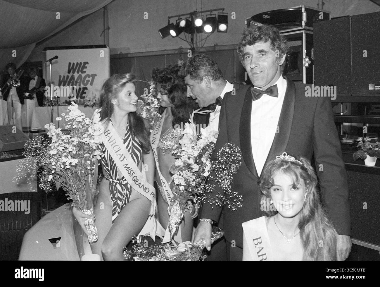 Miss Grand Prix Election Zandvoort, Grand prix, Elections, Zandvoort, 25-08-1984 Whizgle News, Dutch Desk, Paesi Bassi, 1950-2000 Un gruppo di partecipanti al concorso di bellezza, adornato da fasce e mazzi di fiori, si erge insieme in un'atmosfera festosa, mentre un uomo distinto in un smoking si impegna con loro in mezzo a musica e festeggiamenti. Foto Stock