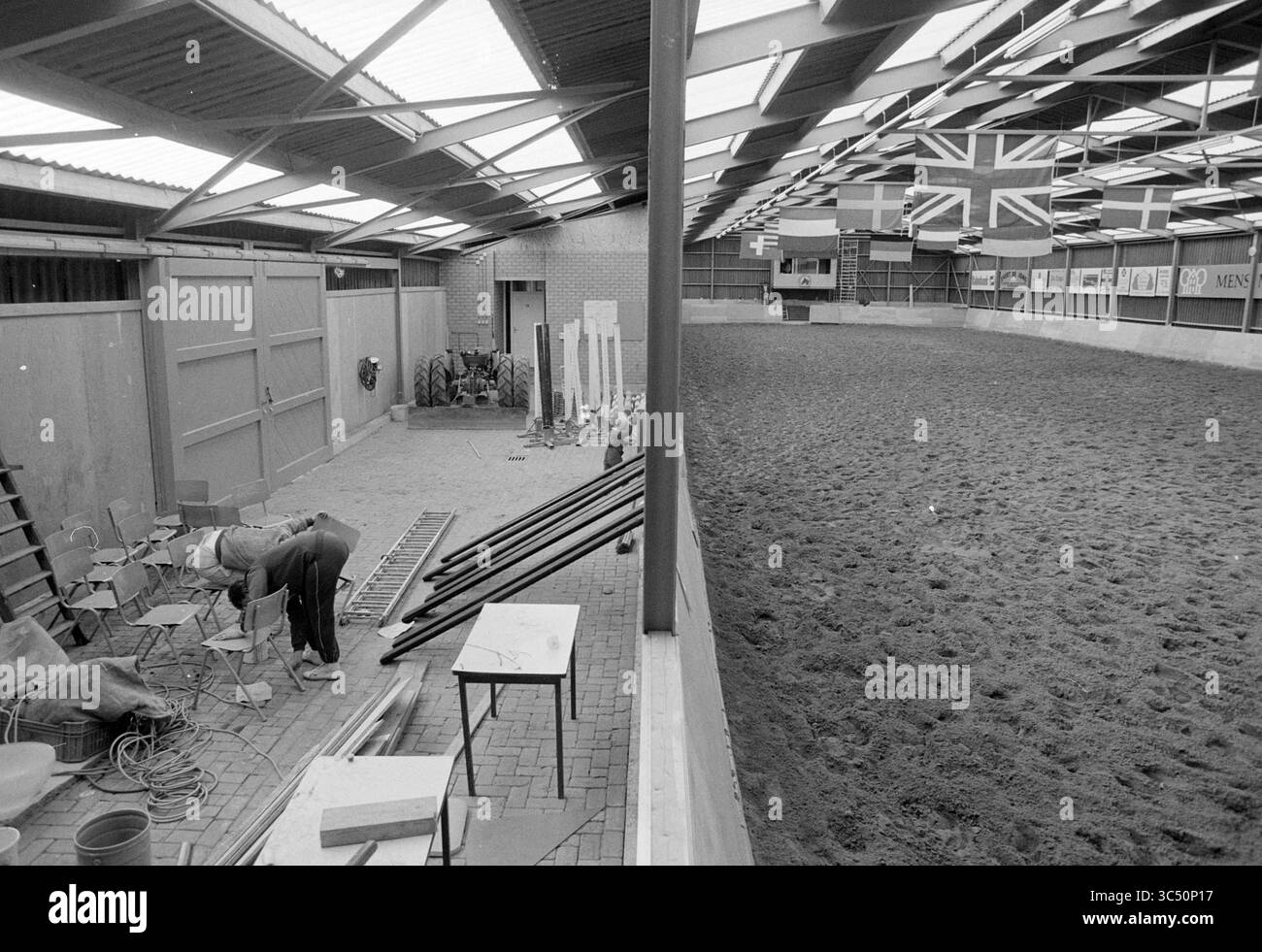 Scuola di equitazione Lisse Roversbroekpolder., scuola di equitazione, Lisse, 21-10-1987 Whizgle News, Dutch Desk, i Paesi Bassi, 1950-2000 un'arena equestre al coperto con pavimento di sabbia circondata da pareti di legno. Varie sedie e attrezzature sono disposte su un lato, mentre bandiere colorate appese al soffitto, aggiungendo un tocco vibrante allo spazio. Foto Stock
