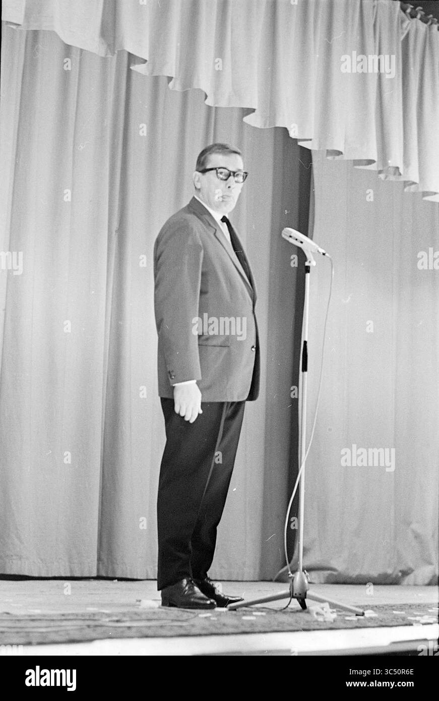 Ria Valk con chitarra Andre Meurs, Cabaret, 13-04-1966 Whizgle News, Dutch Desk, Paesi Bassi, 1950-2000 Un uomo si trova davanti a un microfono sul palco, vestito con un abito con occhiali, rivolgendosi a un pubblico da dietro uno stand. Lo sfondo presenta tende drappeggiate, creando un'atmosfera formale per il suo discorso. Foto Stock