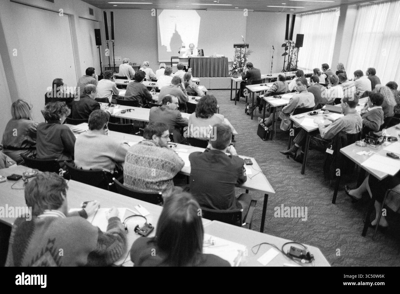 Congress, Damiate, 10-12-1990 Whizgle News, Dutch Desk, Paesi Bassi, 1950-2000 Una sala conferenze piena di partecipanti seduti a lunghi tavoli, impegnati nell'appunti e nell'ascolto di un altoparlante davanti. Un grande schermo visualizza una presentazione, mentre varie apparecchiature audio sono visibili sulle scrivanie. Foto Stock