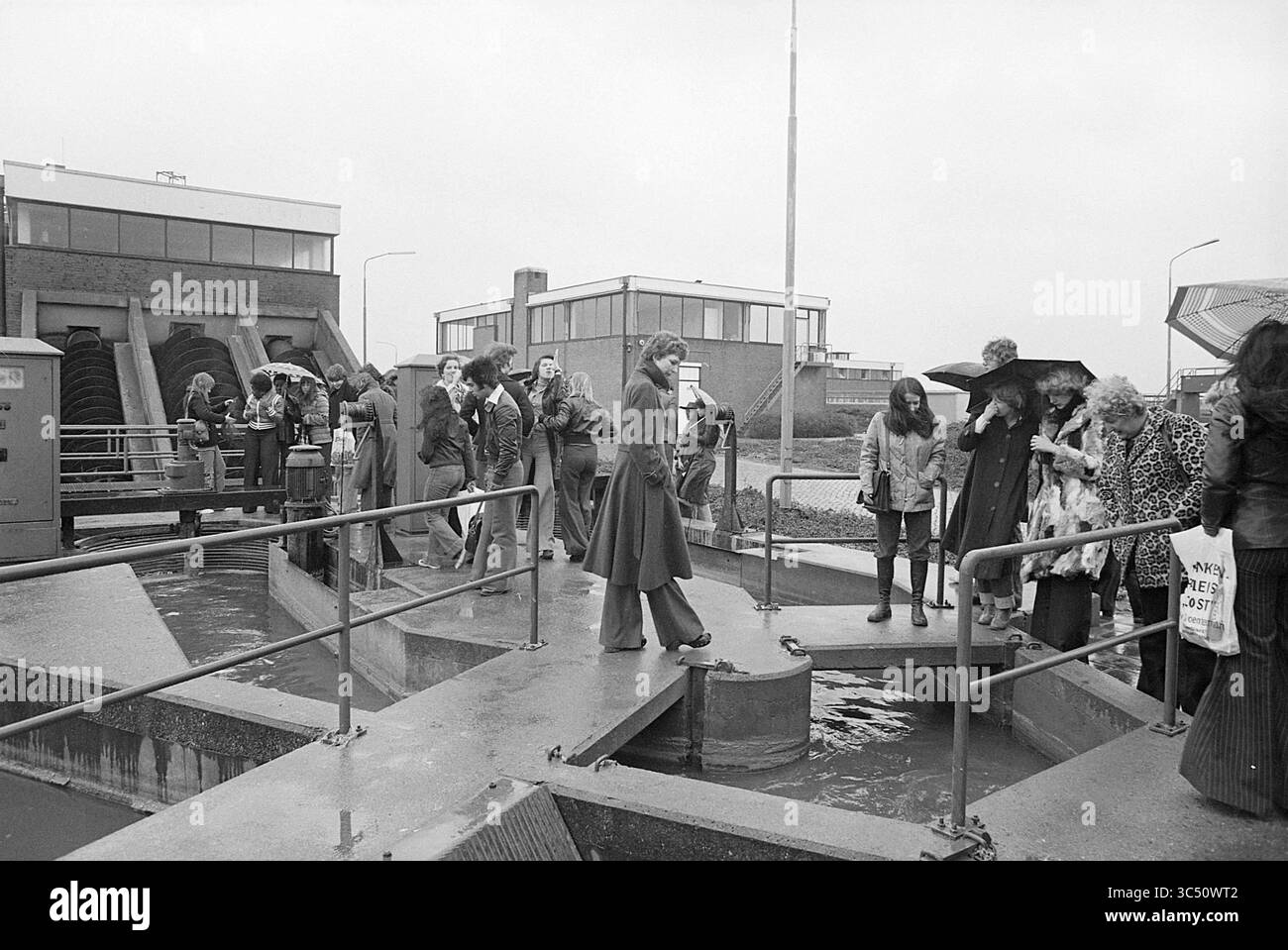 Impianto di trattamento delle acque per escursioni Waarderpolder, escursioni, approvvigionamento idrico, Haarlem, A. Hofmanweg, paesi Bassi, 01-04-1977 Whizgle News, Dutch Desk, Paesi Bassi, 1950-2000 Un gruppo di persone, vestite con abiti diversi, si riunisce intorno a un impianto di trattamento delle acque, impegnato in conversazione e osservazione. Diversi individui tengono gli ombrelli, suggerendo un giorno coperto, mentre altri si trovano su una piattaforma che si affaccia su macchinari e acqua. Alcuni partecipanti mostrano dei cartelli che indicano una dimostrazione o un evento di sensibilizzazione ambientale. Foto Stock