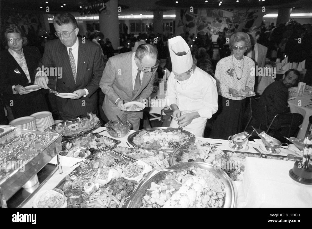 Hoogovens Anniversary Meeting the Hague, Hoogovens, Den Haag, 21-11-1989 Whizgle News, Dutch Desk, i Paesi Bassi, 1950-2000 Un vivace scenario di banchetti con una varietà di piatti presentati elegantemente, mentre gli ospiti si servono con entusiasmo mentre uno chef prepara con cura il cibo. L'atmosfera e' vivace, con i partecipanti impegnati in conversazione e gustando le offerte culinarie. Foto Stock