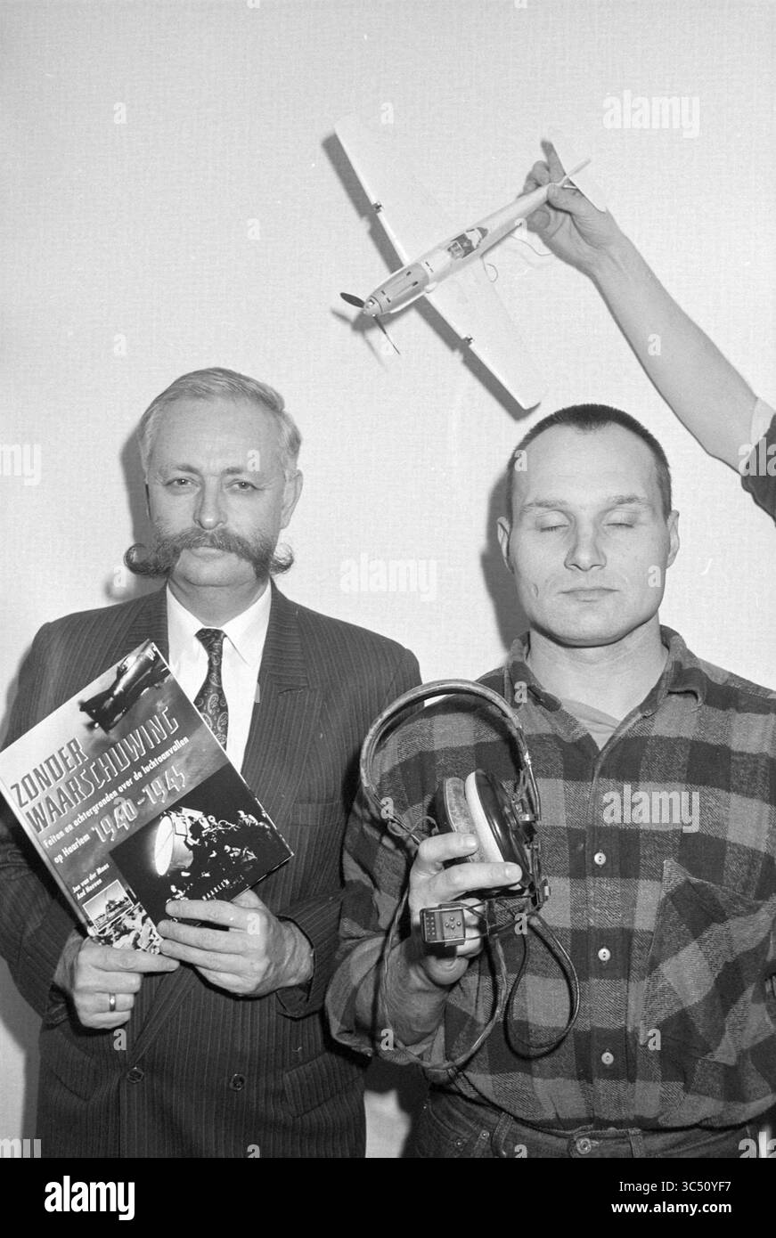 Va bene senza preavviso, 14-02-1995 Whizgle News, Dutch Desk, Paesi Bassi, 1950-2000 due uomini stanno fianco a fianco, uno indossa un abito e tiene in mano un libro intitolato "Zonder Waarschuwing", mentre l'altro, vestito in plaid, tiene una serie di cuffie. Sopra di loro, si vede una mano che solleva in aria un modellino di aeroplano, creando un intrigante contrasto tra le espressioni serie dei due uomini e l'azione stravagante sopra. Foto Stock