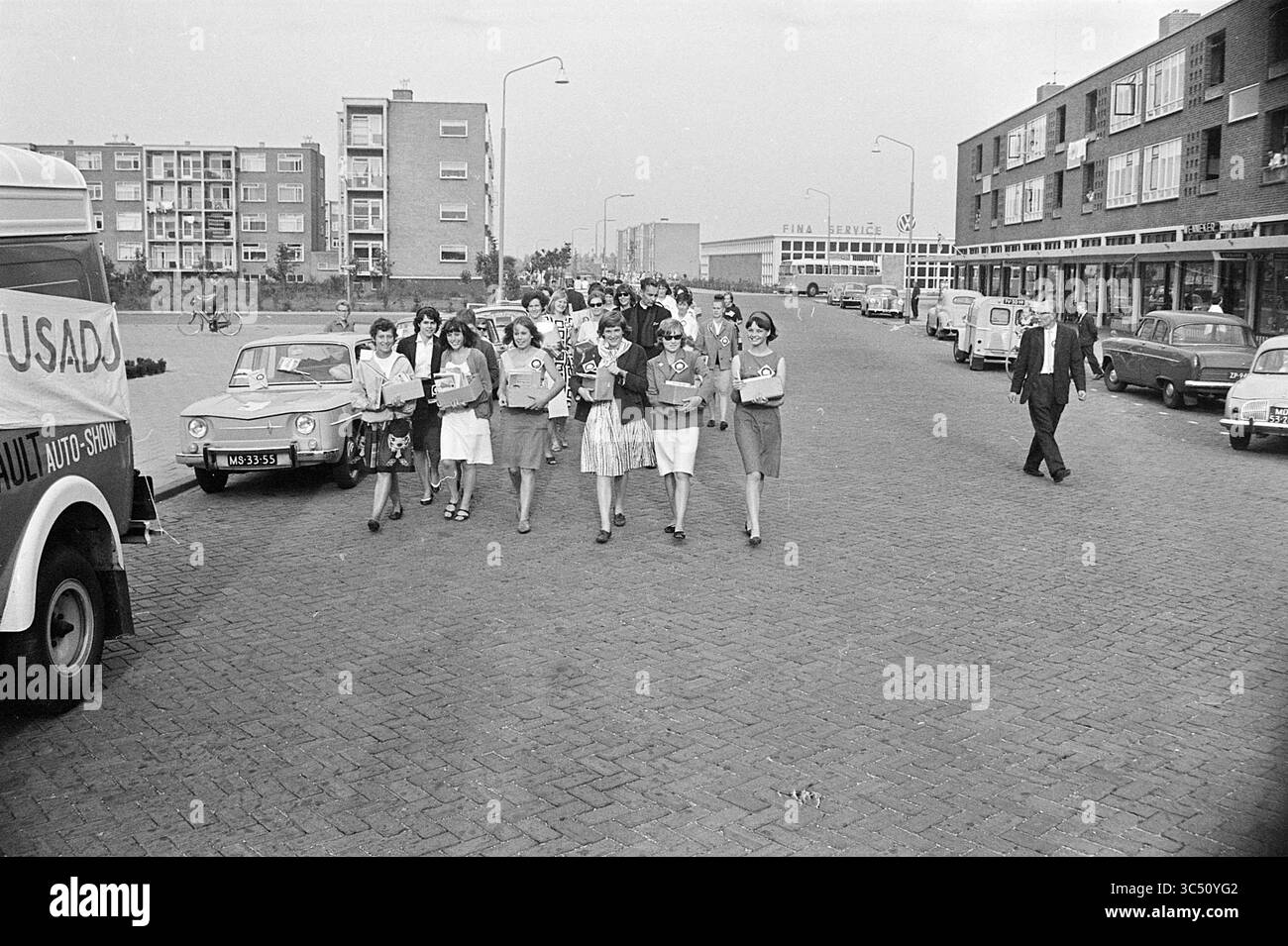 Action Customs (iran), 00-00-1963 Whizgle News, Dutch Desk, Paesi Bassi, 1950-2000 Un gruppo di giovani donne in abiti eleganti cammina insieme lungo una strada lastricata, portando libri e documenti. Sembrano far parte di un evento o di un rally organizzato, con diversi veicoli retrò parcheggiati nelle vicinanze e edifici caratteristici che fiancheggiano la strada. Alcuni individui, tra cui un uomo solitario, possono essere visti impegnati in varie attività intorno a loro. Foto Stock