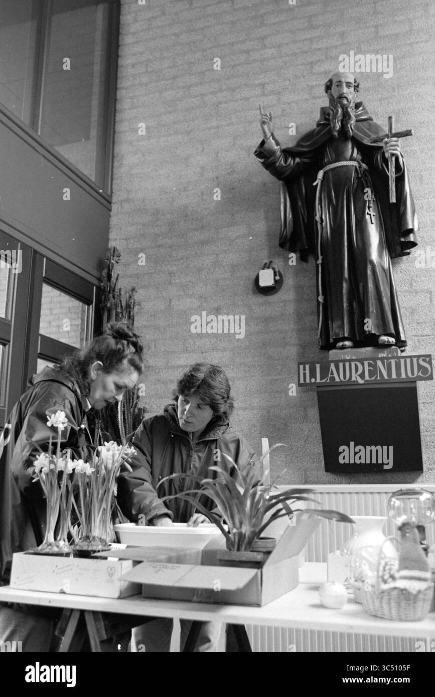Mercato delle pulci, Fidelishof, IJm., IJmuiden, Paesi Bassi, 07-03-1997 Whizgle News, Dutch Desk, Paesi Bassi, 1950-2000 due persone sono impegnate in un'attività a un tavolo decorato con piante in vaso, mentre una statua di un santo li sorveglia dal muro sopra, contribuendo a un'atmosfera serena e riflettente. Foto Stock