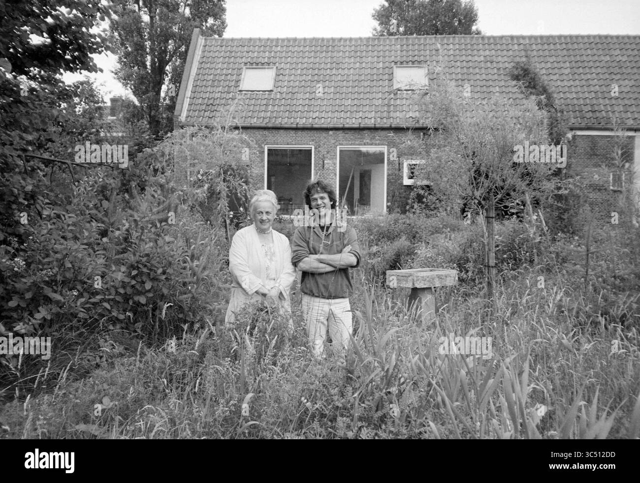 Report Heemstede, Heemstede, Paesi Bassi, 18-07-1984 Whizgle News, Dutch Desk, i Paesi Bassi, 1950-2000 due persone stanno insieme in un giardino lussureggiante, sorridente tra erba alta e arbusti, con una casa accogliente sullo sfondo. La scena trasmette un senso di cameratismo e connessione con la natura. Foto Stock