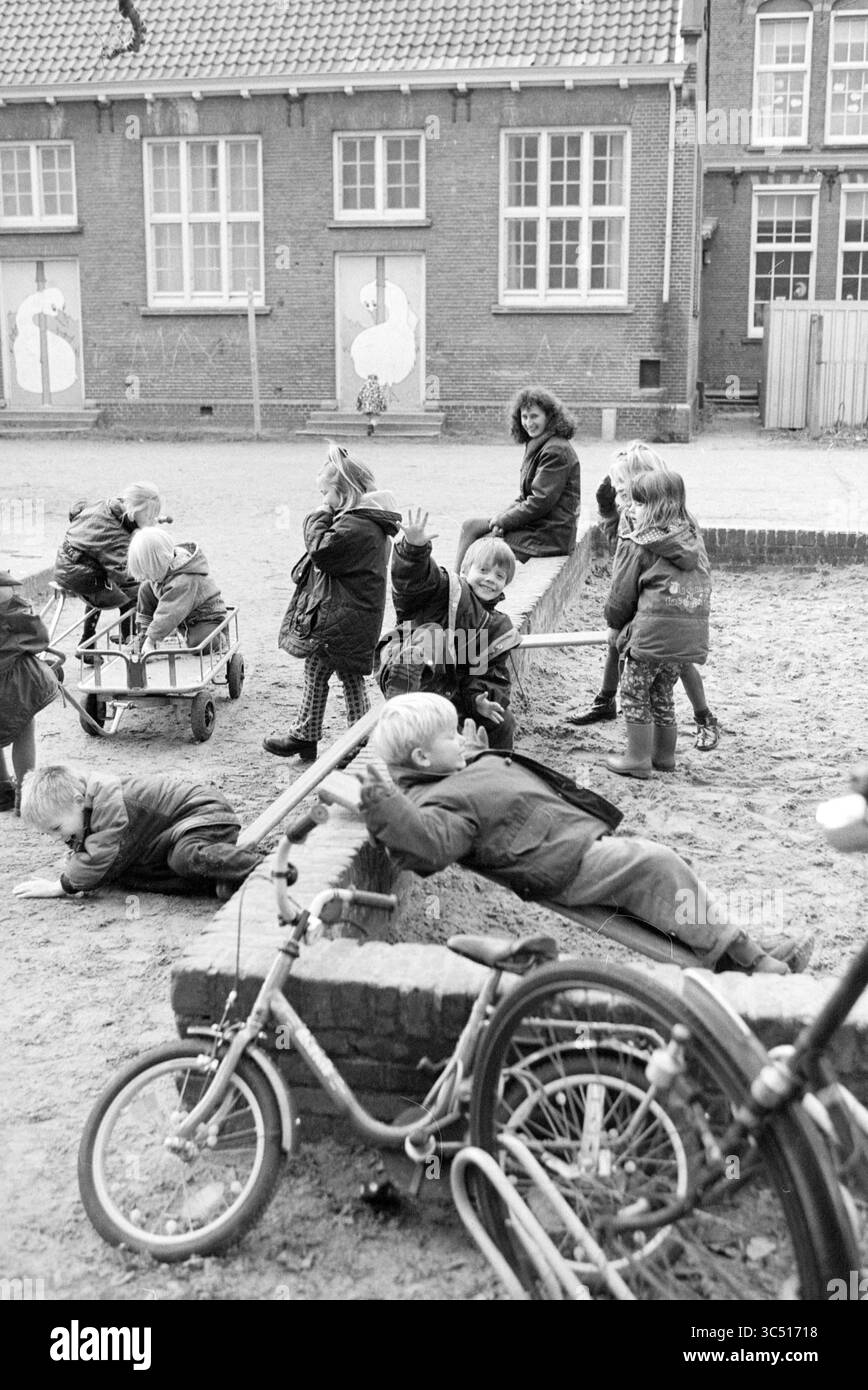 Children on School Playground, 16-11-1998 Whizgle News, Dutch Desk, The Netherlands, 1950-2000 Un gruppo di bambini gioca in un'area sabbiosa, impegnandosi in varie attività. Alcuni stanno sdraiandosi su una parete bassa, altri spingono i carri giocattolo, e alcuni sono in bicicletta. Una donna li guarda con un sorriso, creando una vivace scena di gioia giovanile ed esplorazione. Gli edifici con grandi finestre fungono da sfondo, accennando a una scuola o a uno spazio comune nelle vicinanze. Foto Stock