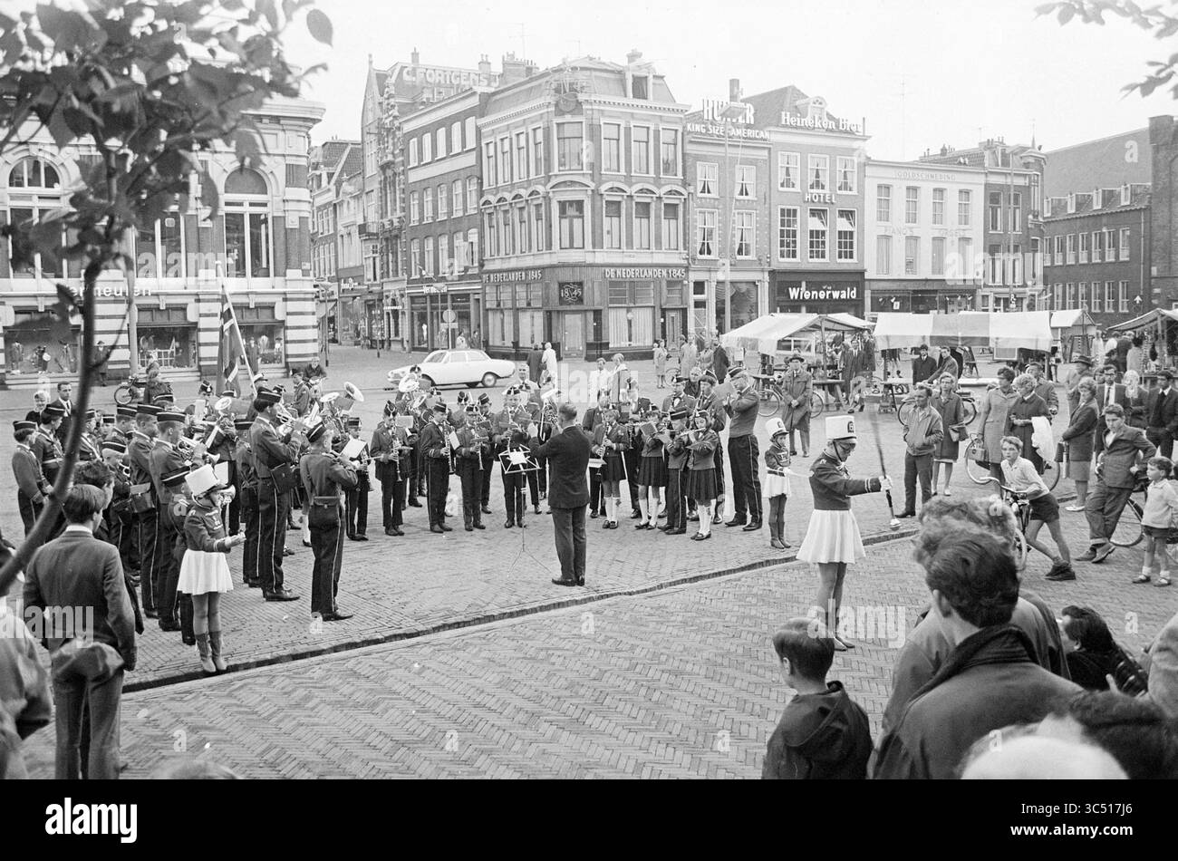 Gruppo norvegese Grote Markt, Grote Markt, musica, Haarlem, Grote Markt, The Netherlands, 28-06-1967','29-06-1967 Whizgle News, Dutch Desk, The Netherlands, 1950-2000 Un vivace raduno in una piazza della città, con una banda in marcia che si esibisce di fronte a una folla. Persone di tutte le età guardano con attenzione, mentre alcune scattano foto. Gli edifici circostanti mostrano un mix di vetrine e segni, creando un vibrante sfondo urbano. Bandiere e striscioni aggiungono un'atmosfera festosa alla scena. Foto Stock