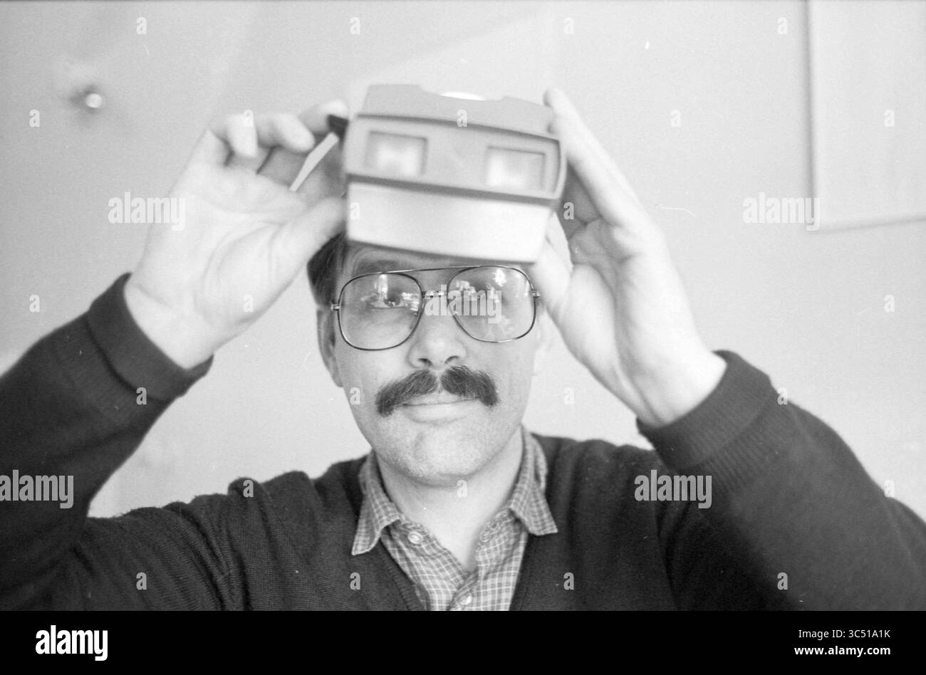 Jan Zumbrink, art editor, Haarlems Dagblad ecc. e IJmuider Courant e Koeri, 01-11-1987 Whizgle News, Dutch Desk, Paesi Bassi, 1950-2000 Un uomo che indossa grandi occhiali e baffi tiene un mirino vintage sopra gli occhi, sorridendo dolcemente. Indossa un maglione sopra una camicia con colletto e lo sfondo suggerisce un ambiente interno accogliente. Foto Stock