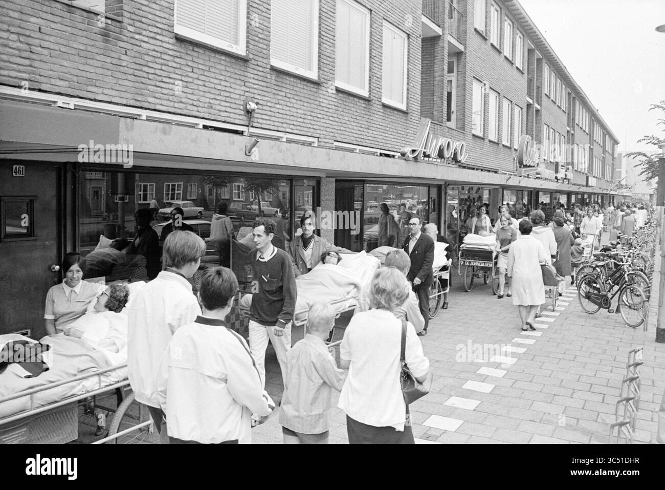 I malati hanno guidato lungo Lange Nieuwstraat, Sick and Hospitals, IJmuiden, Lange Nieuwstraat, Paesi Bassi, 03-07-1969 Whizgle News, Dutch Desk, Paesi Bassi, 1950-2000 Una scena di strada trafficata con un gruppo di persone, alcuni che spingono letti di ospedale con i pazienti, mentre altri aspettano o interagiscono nelle vicinanze, accanto a una fila di edifici e biciclette parcheggiate. L'atmosfera suggerisce un ambiente medico o sanitario, con un mix di personale e visitatori impegnati in varie attività. Foto Stock