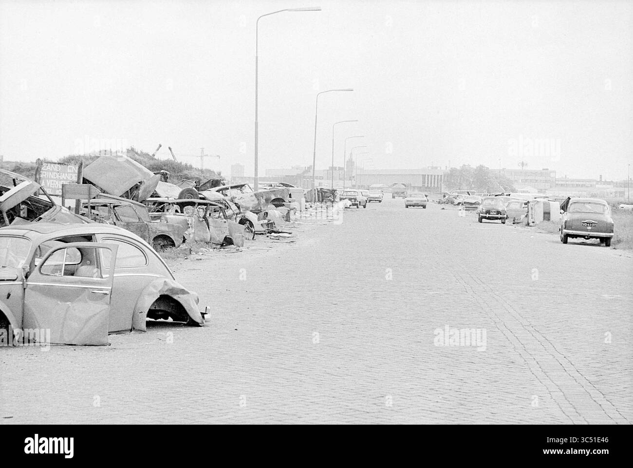 Rottami del cimitero automobilistico, 18-06-1971 Whizgle News, Dutch Desk, Paesi Bassi, 1950-2000 una strada abbandonata costeggiata da auto abbandonate, alcune parzialmente smantellate, crea una scena inquietante di abbandono e degrado. La vegetazione ricoperta e le strutture lontane suggeriscono un'area un tempo trafficata ora lasciata al tempo. Foto Stock