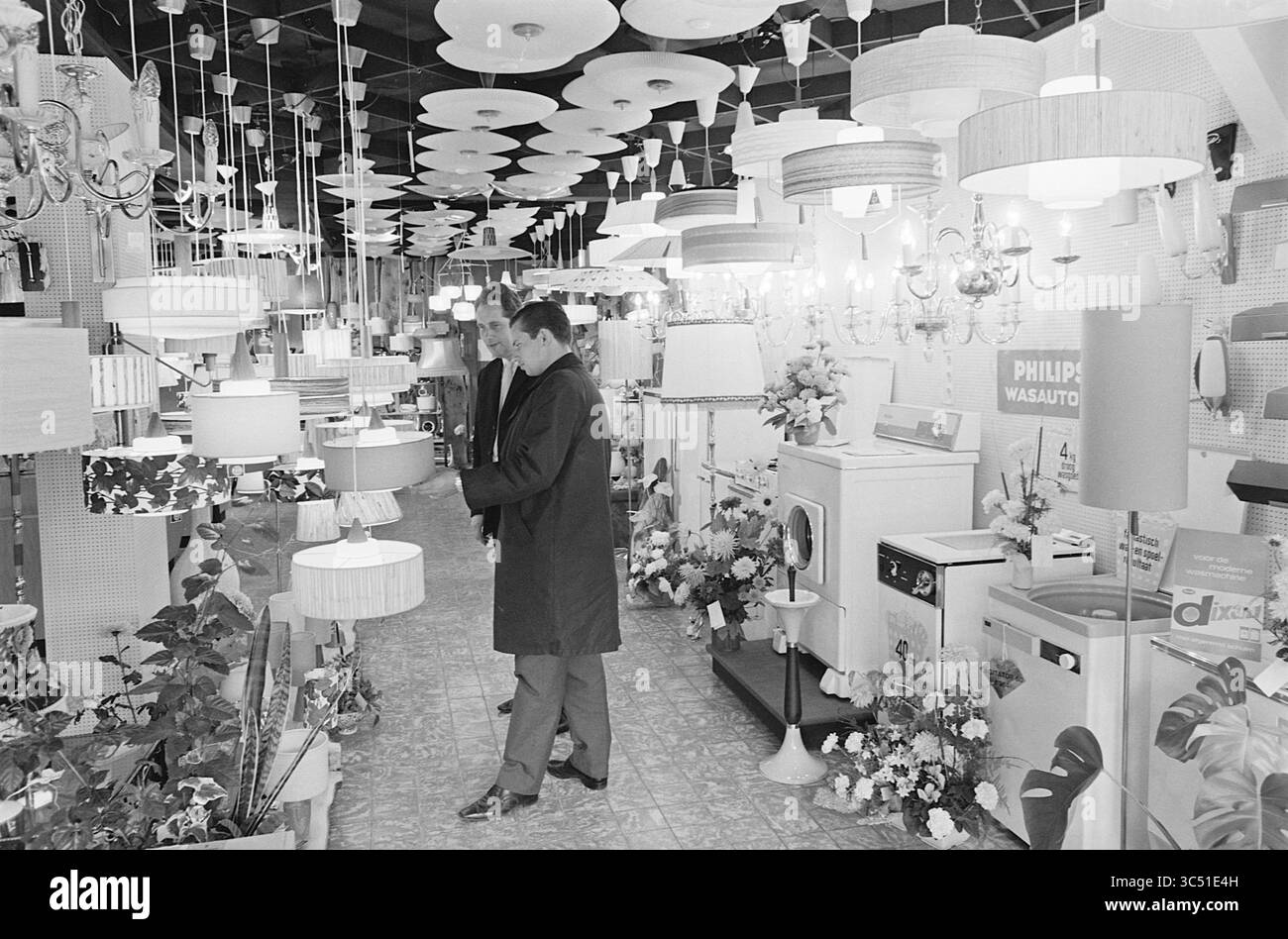 Lighting Rigo, Interior, 18-09-1964 Whizgle News, Dutch Desk, Paesi Bassi, 1950-2000 due persone esplorano uno showroom ben illuminato con una serie di lampade a sospensione e lampade, circondato da piante decorative e composizioni floreali, che mostrano vari design e stili di prodotti di illuminazione. Foto Stock Lighting Rigo, Interior, 18-09-1964 Whizgle News, Dutch Desk, Paesi Bassi, 1950-2000 due persone esplorano uno showroom ben illuminato con una serie di lampade a sospensione e lampade, circondato da piante decorative e composizioni floreali, che mostrano vari design e stili di prodotti di illuminazione. Foto Stock