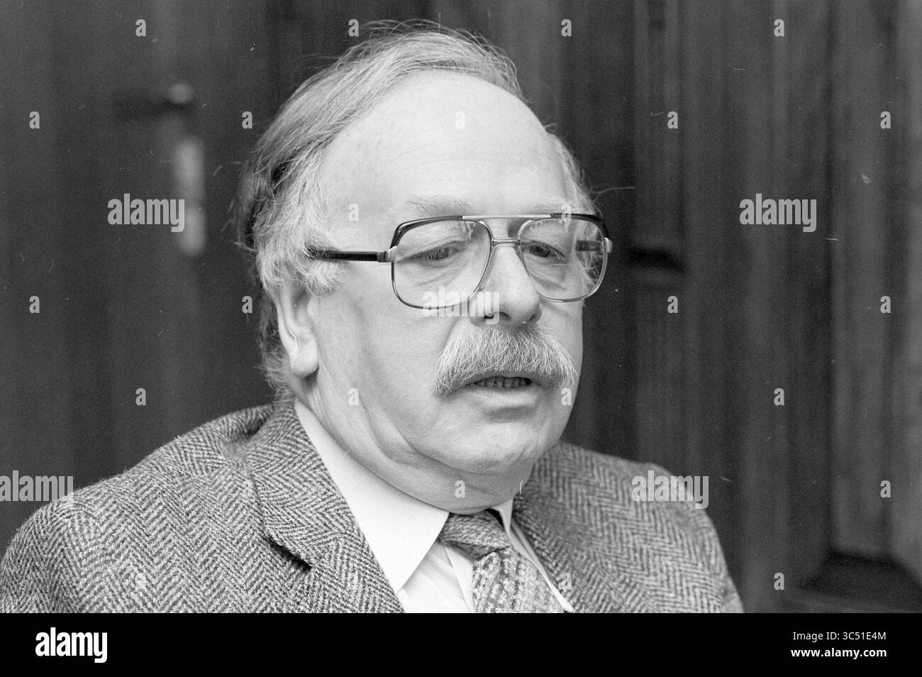 Sig. Rijkers Alderman Beverwijk, persone, Aldermen, Beverwijk, Paesi Bassi, 31-01-1984 Whizgle News, Dutch Desk, Paesi Bassi, 1950-2000 un uomo più anziano con occhiali e baffi siede con cura, indossando una giacca in tweed. La sua espressione suggerisce la contemplazione, con uno sfondo di texture di legno che aggiungono alla serietà della scena. Foto Stock