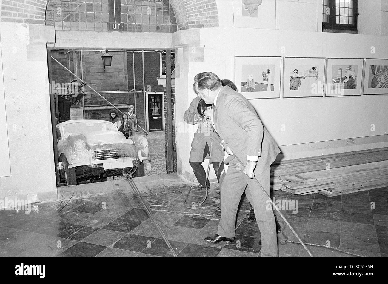 Building the Exhibition Vleeshal, Exhibitions, 09-02-1971 Whizgle News, Dutch Desk, Paesi Bassi, 1950-2000 Un gruppo di uomini si vede tirare un'auto d'epoca attraverso una porta aperta, con opere d'arte appese alle pareti nelle vicinanze. La scena cattura un mix di installazione artistica urbana e ingegneria inaspettata, mettendo in risalto un impegno creativo in uno spazio pubblico. Foto Stock