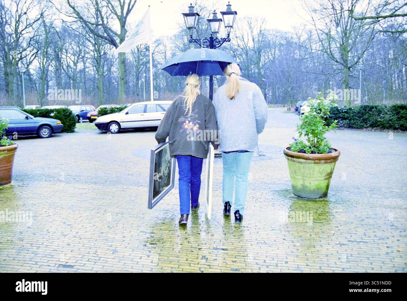 Valuation by Christie's in Heemstede, Heemstede, Paesi Bassi, 30-01-1995 Whizgle News, Dutch Desk, i Paesi Bassi, 1950-2000 due persone camminano mano nella mano, condividendo un ombrello mentre trasportano una foto incorniciata. Navigano in un'area esterna piovosa, circondata da alberi e auto parcheggiate, creando un'atmosfera serena. Foto Stock